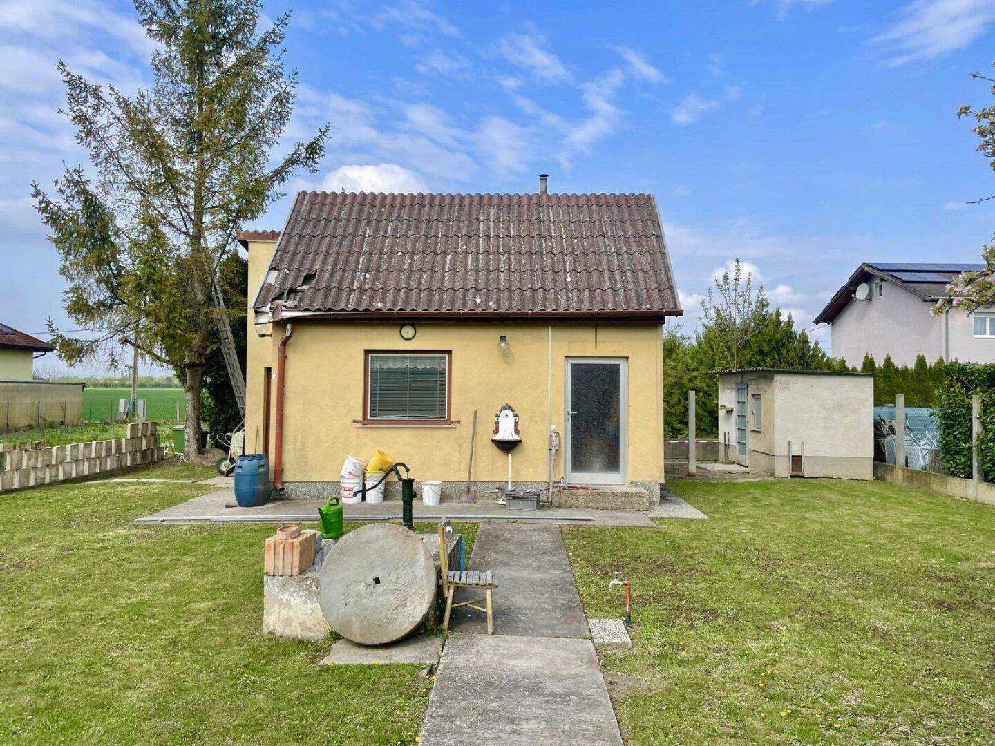 Vorderansicht des gelben Hauses mit Ziegeldach, Garten und einer Wasserpumpe.