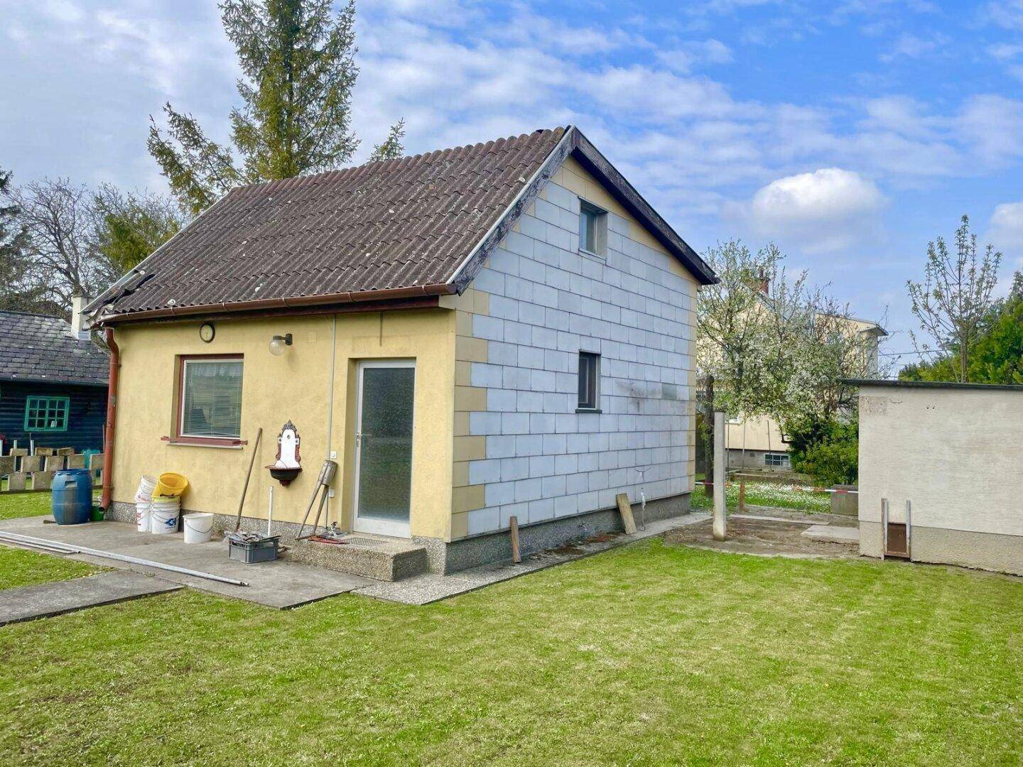 Seitenansicht des Hauses mit gelber Fassade und teilweise verkleideter Wand, umgeben von Rasen.