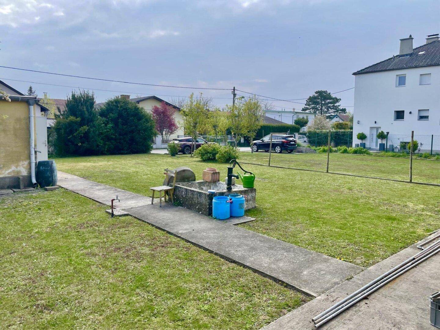 Weitläufiger Garten mit Betonweg, Wasserpumpe und Blick auf die Nachbarschaft.