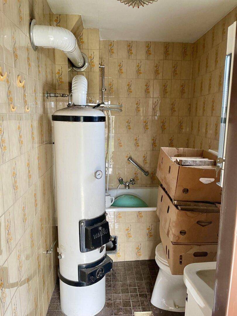 Badezimmer mit beigen Fliesen, Badewanne, Toilette und einem großen Warmwasserboiler.