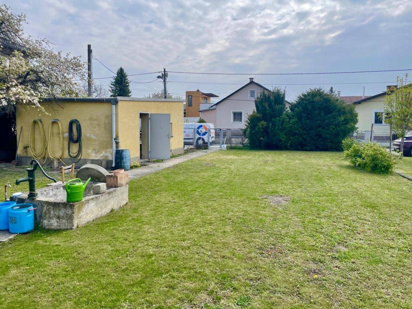 Gartenbereich mit einem gelben Schuppen, einer Wasserpumpe und weitem Rasen.