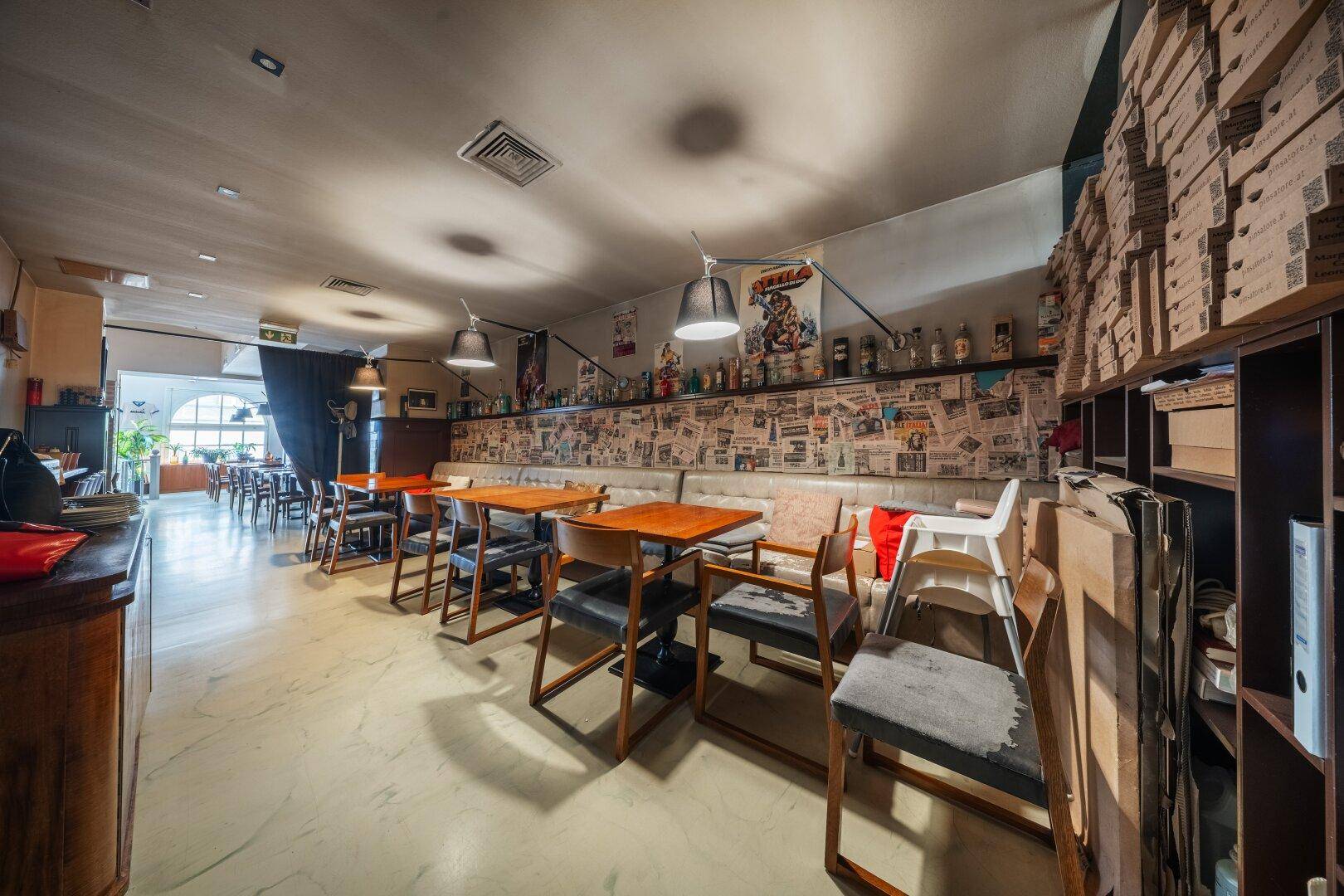 Langer Gastraum mit Sitzbänken, Holztischen und Regalen voller Kartons an der Wand.