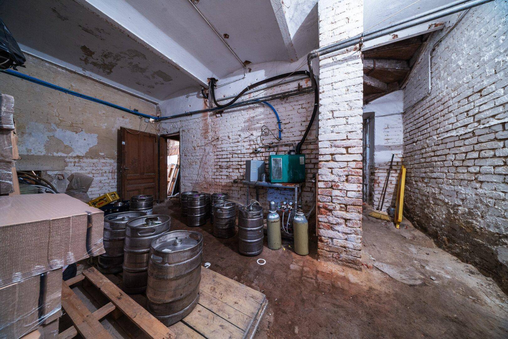 Rustikaler Kellerraum mit unverputzten Ziegelwänden, Bierfässern und sichtbaren Leitungen.