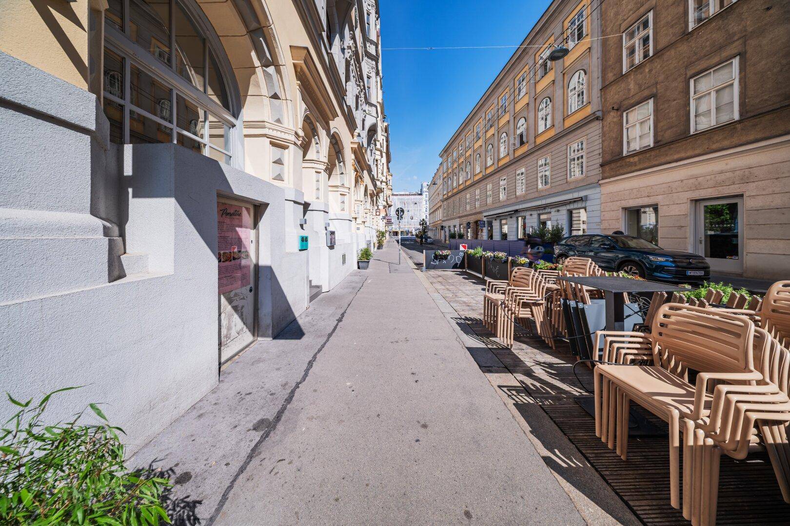 Ruhige Stadtstraße mit Gebäudefassaden, Bürgersteig und einer Außenterrasse mit Stühlen.