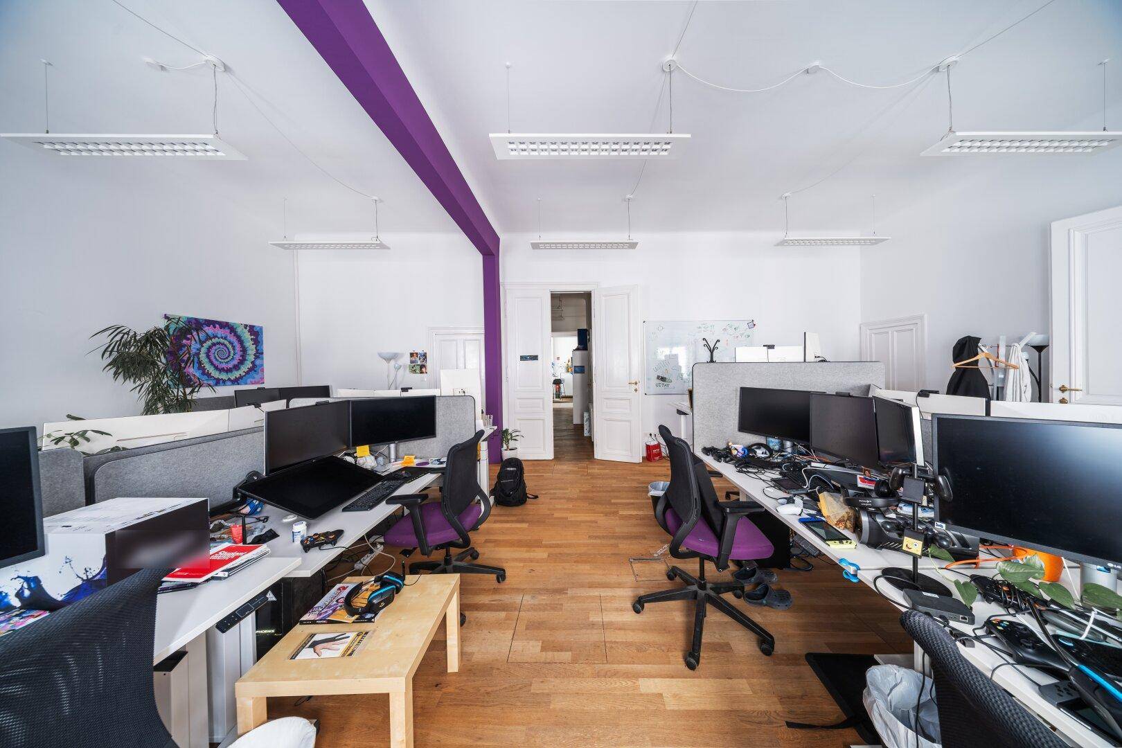 Großes Büro mit Holzboden, vielen Arbeitsplätzen und einer lila Akzentwand, die den Raum strukturiert.