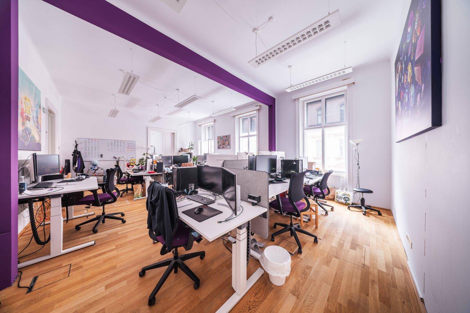 Weitläufiges Büro mit Holzboden, vielen Arbeitsplätzen und einer durchgehenden lila Akzentwand.