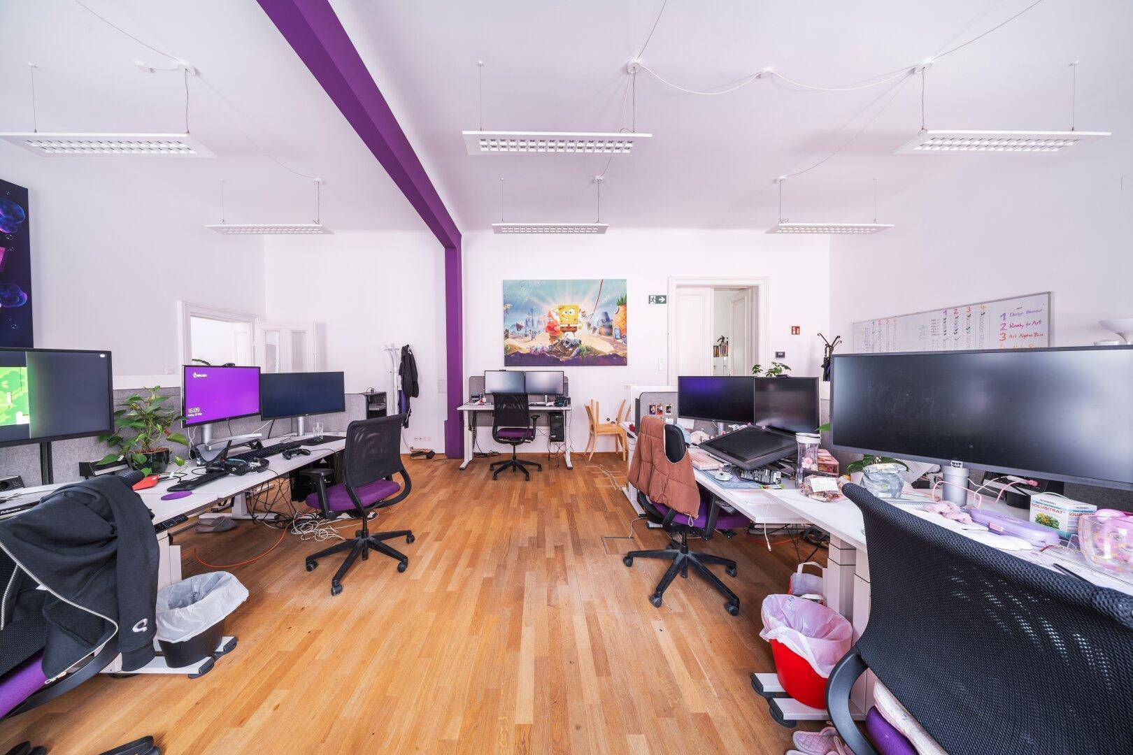 Helles Büro mit Holzboden, mehreren Arbeitsplätzen und einer lila Akzentwand, die den Raum belebt.