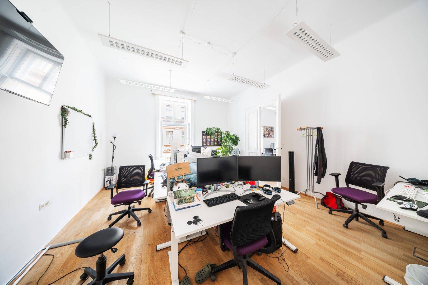 Offenes Büro mit Holzboden, mehreren Arbeitsplätzen und einem großen Fenster, das den Raum erhellt.