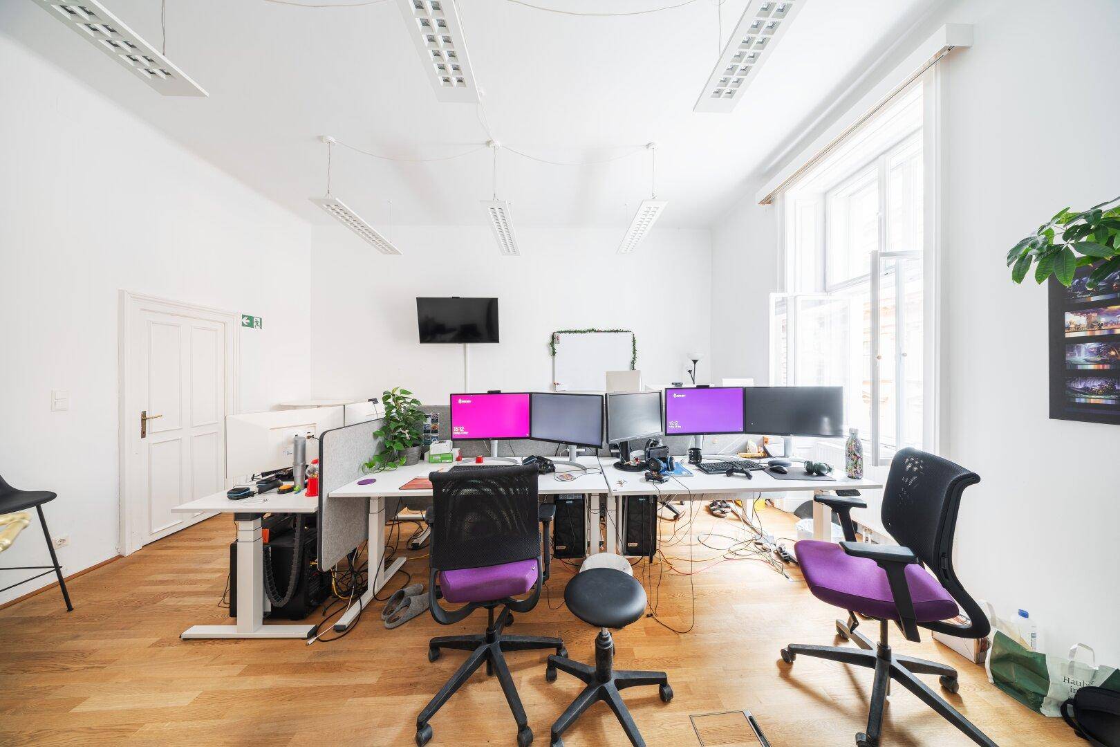 Modernes Büro mit Holzboden, mehreren Arbeitsplätzen und großen Fenstern für viel Tageslicht.