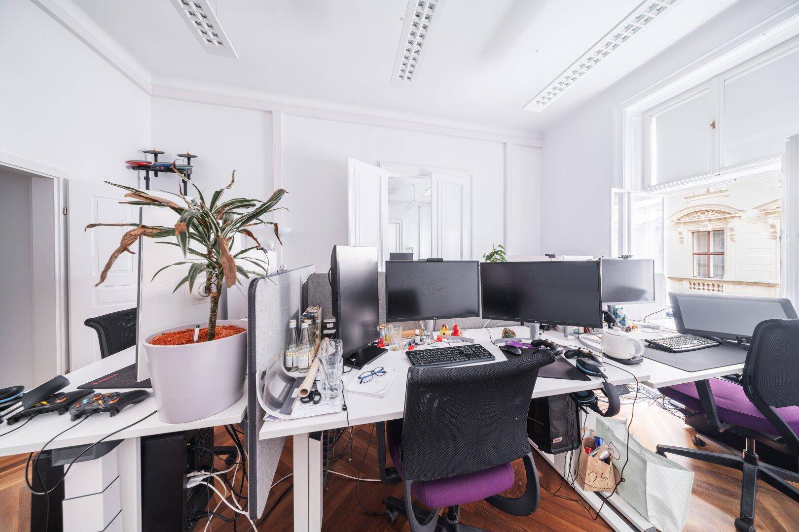 Detailansicht eines Arbeitsplatzes in einem offenen Büro mit mehreren Monitoren und Pflanzen.