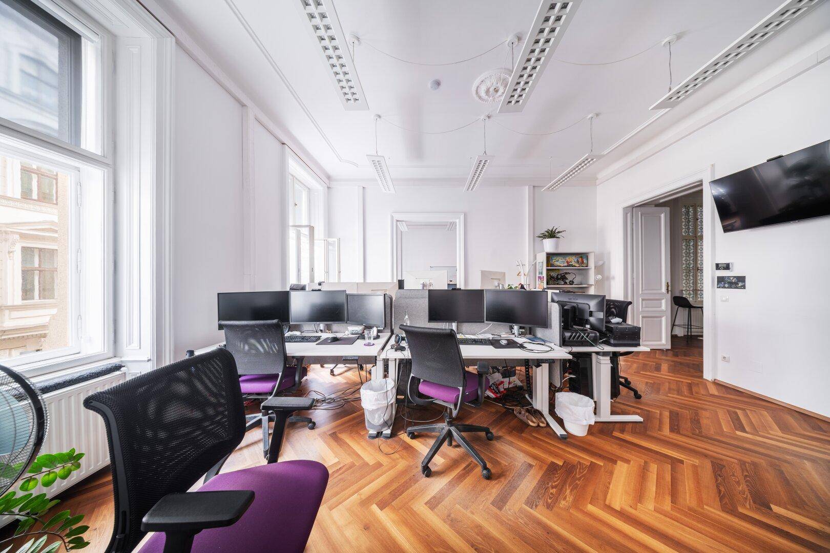 Offener Bürobereich mit mehreren Arbeitsplätzen, Fischgrätparkett und großen Fenstern.
