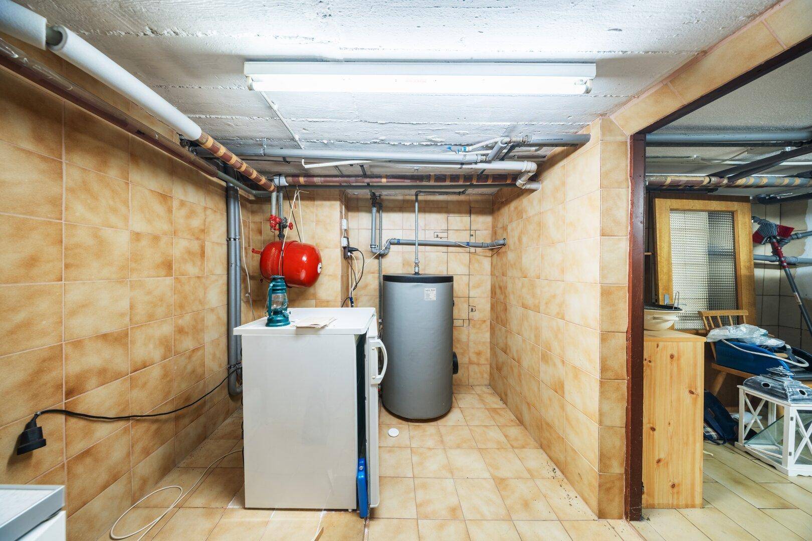 Gefliester Kellerraum mit Warmwasserboiler, Leitungen und einem Kühlschrank.