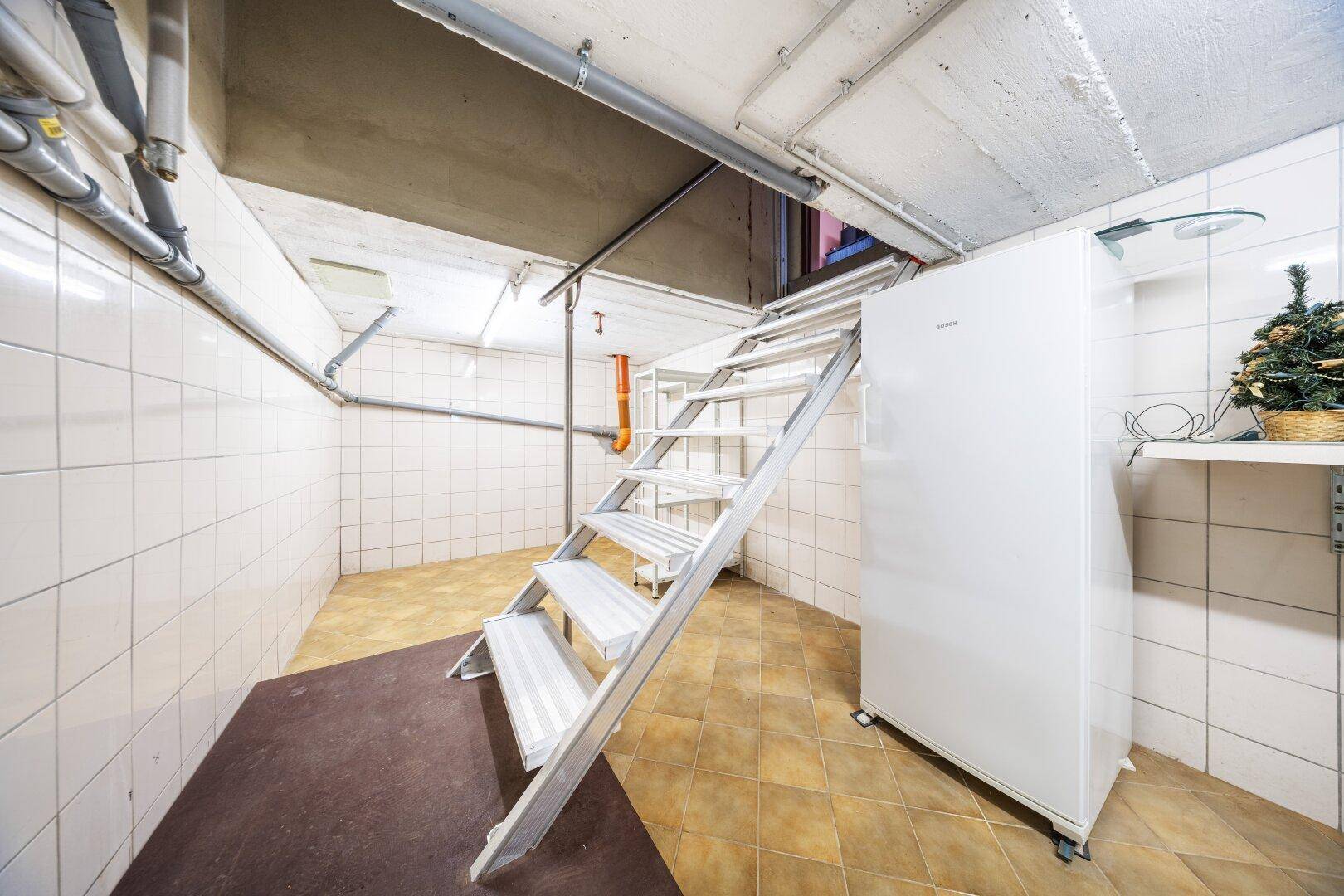 Kellerraum mit Metalltreppe, Kühlschrank und gefliesten Wänden.