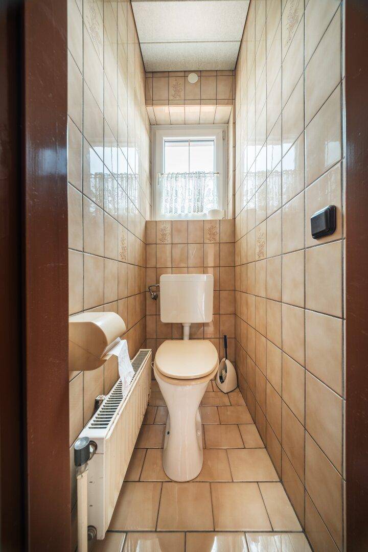 Kleines, gefliestes WC mit Fenster, Heizkörper und einfacher Toilette.