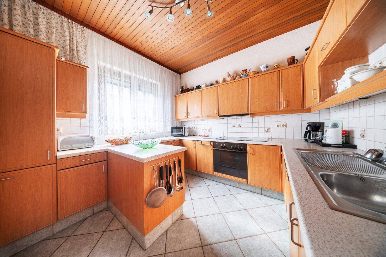 Küche mit Holzschränken, Fliesenboden und Fenster mit viel Tageslicht.