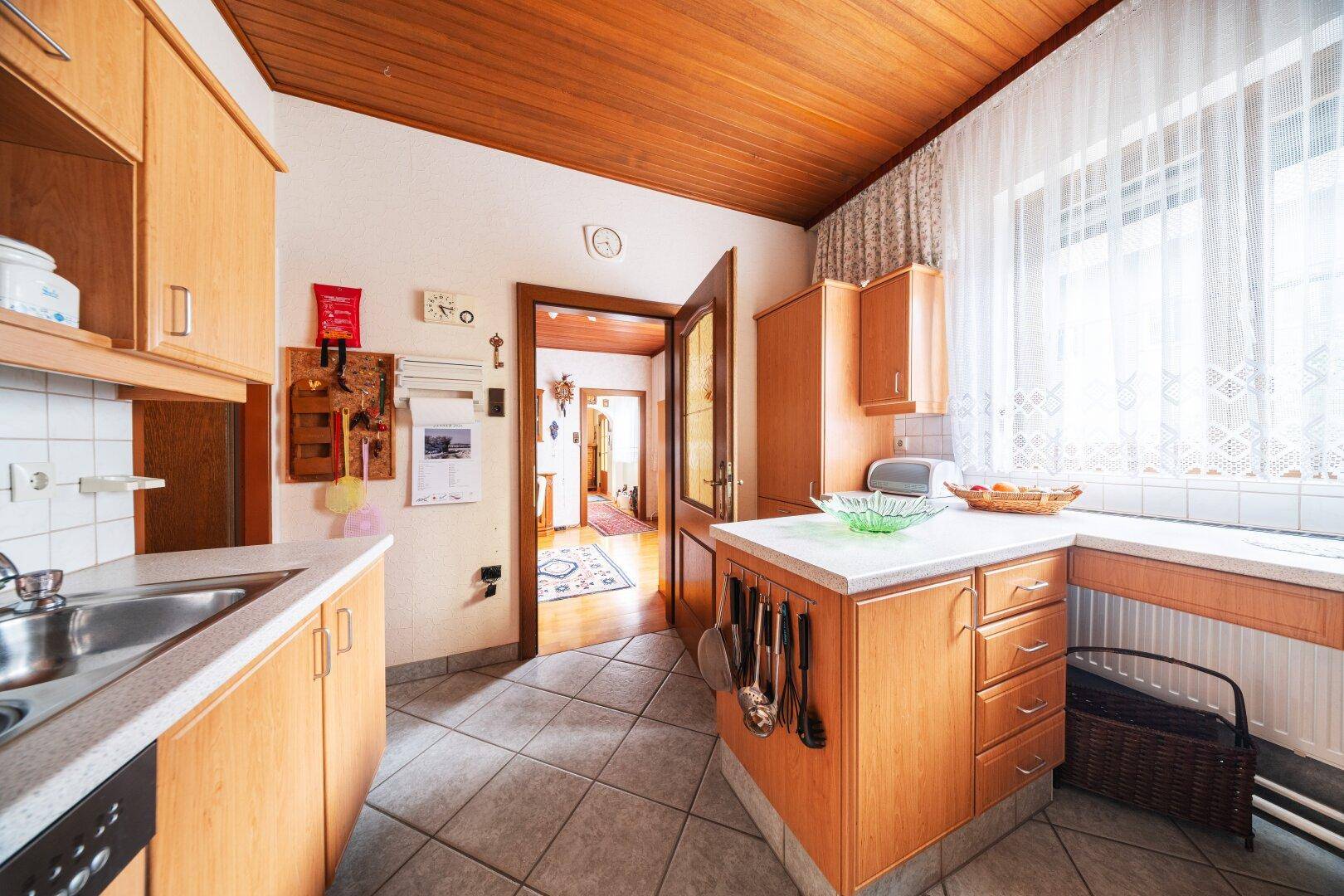 Küche mit Holzschränken, Fliesenboden und Fenster mit viel Tageslicht.