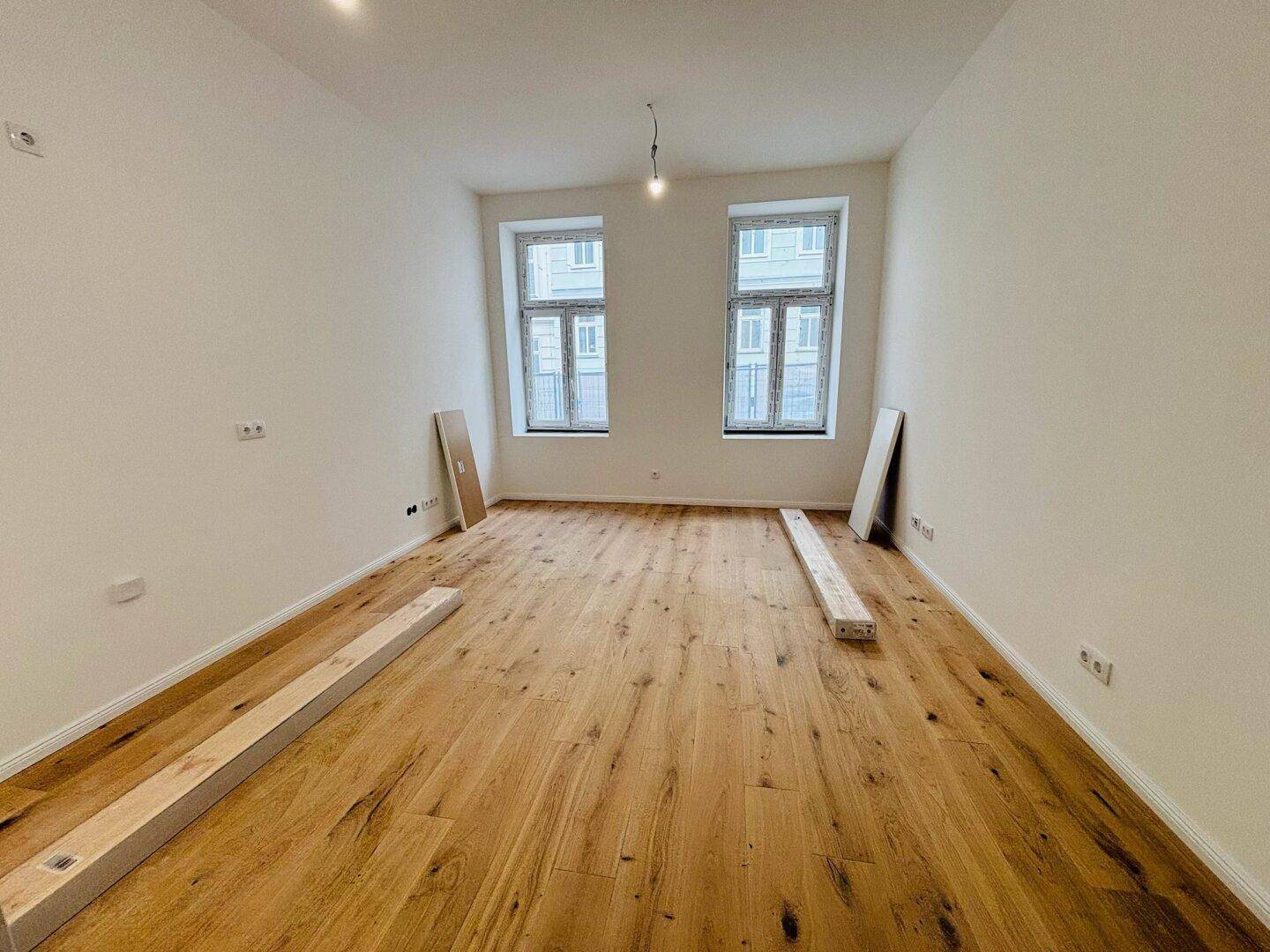 Leeres Wohnzimmer mit neuem Holzboden, hohen Decken und großen Fenstern.