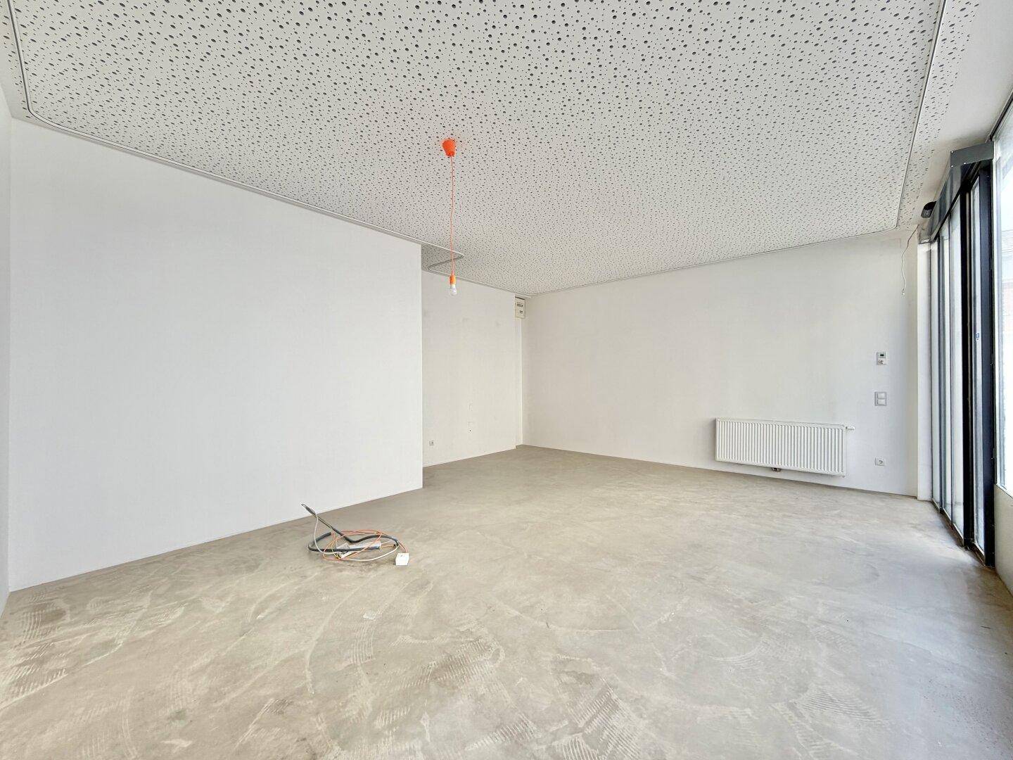 Leerer Gewerberaum mit Heizkörper an der Wand, großen Fenstern und einem schlichten Betonboden.