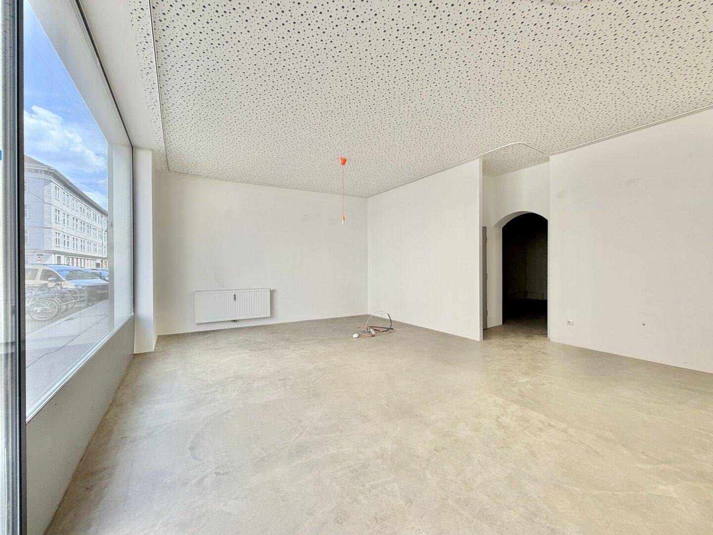 Geräumiger Geschäftsraum mit großen Fenstern, Betonboden und einem Durchgang zu weiteren Bereichen.