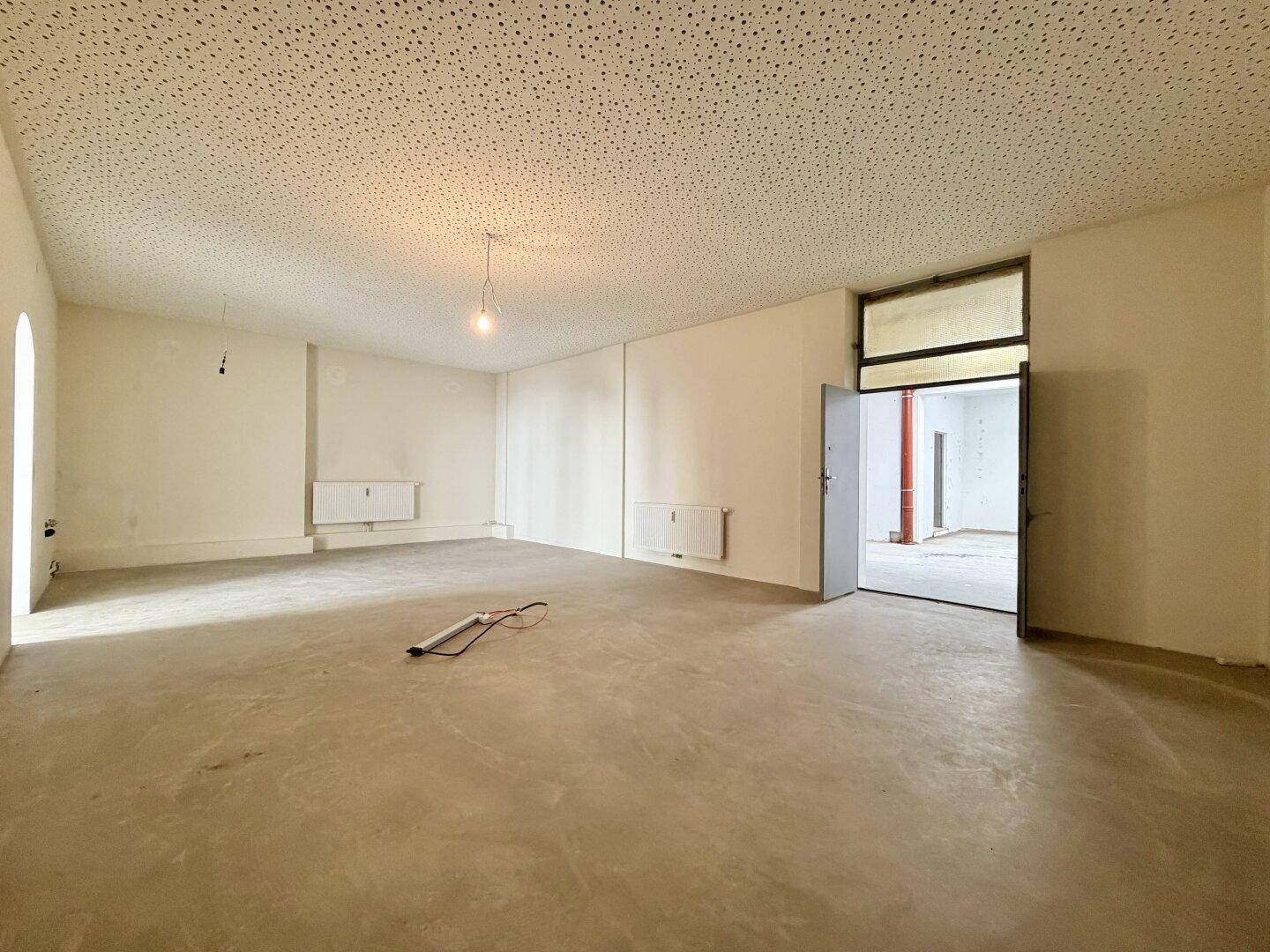 Heller Gewerberaum mit Betonboden, Heizkörper und offener Tür zu einem angrenzenden Bereich.