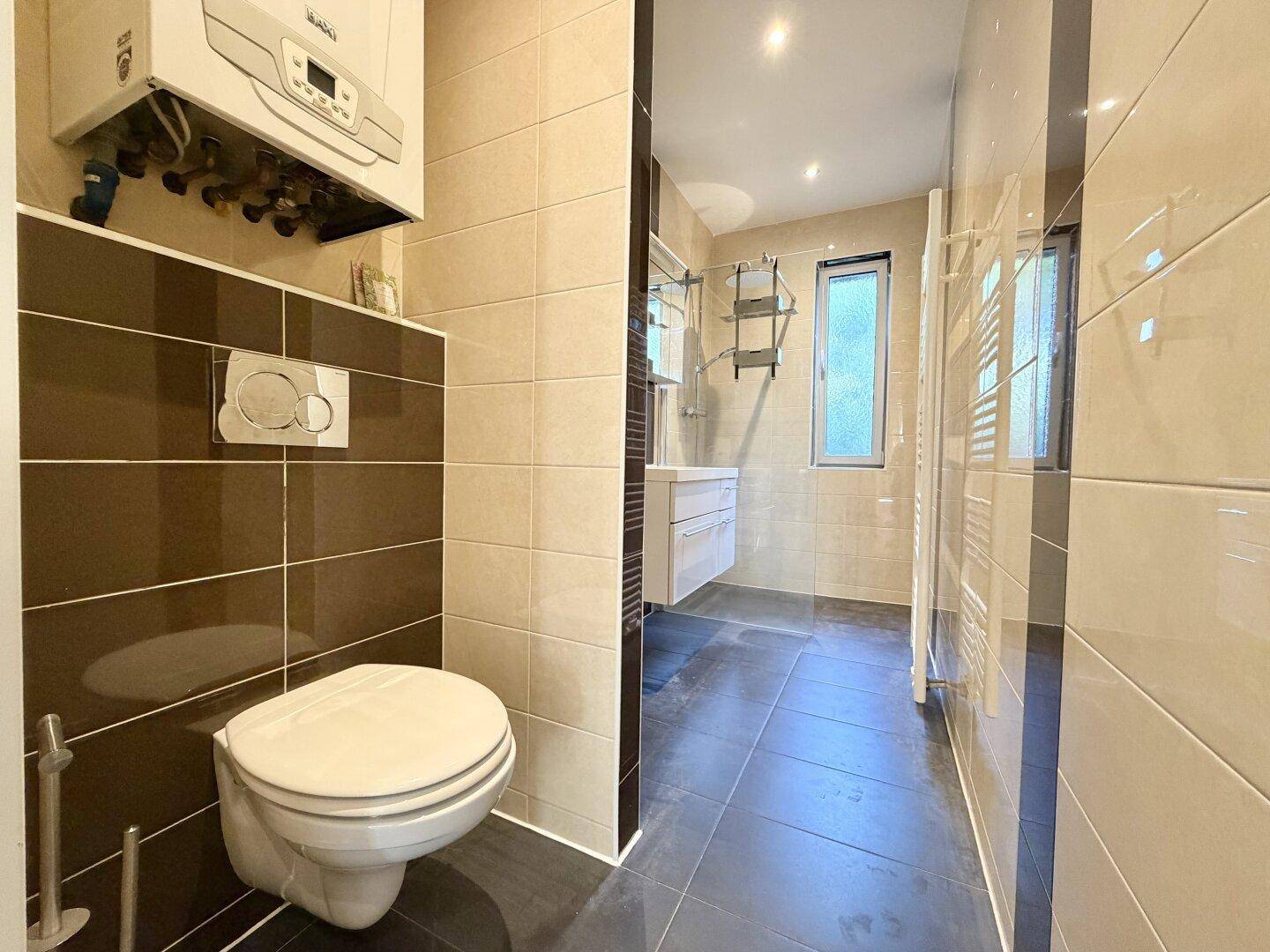 Badezimmer mit Wand-WC, beigen und braunen Fliesen und Heizung, effiziente Raumnutzung.