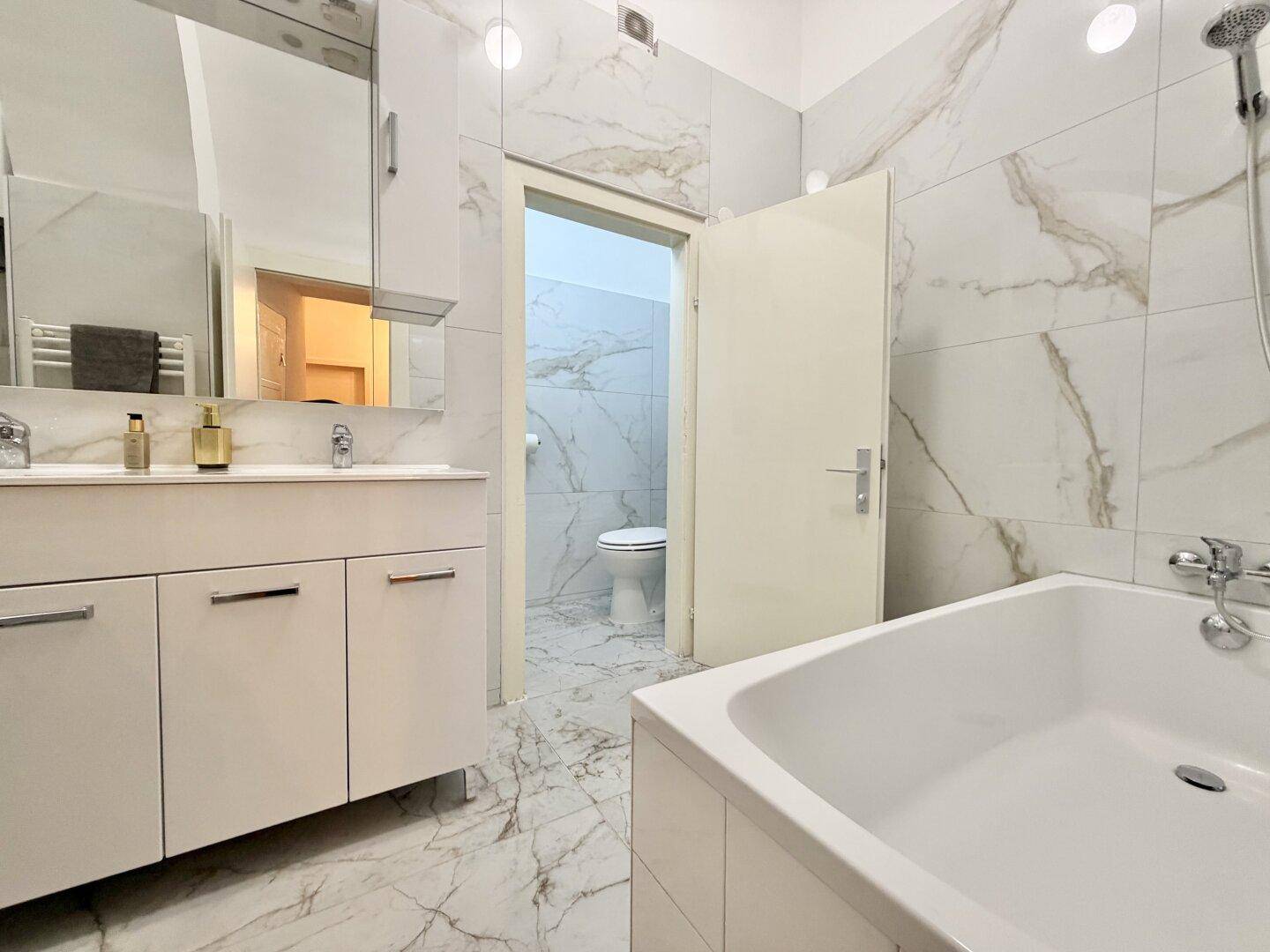 Helles Badezimmer mit Badewanne und Zugang zu einer separaten Toilette, alles in Marmoroptik gefliest.