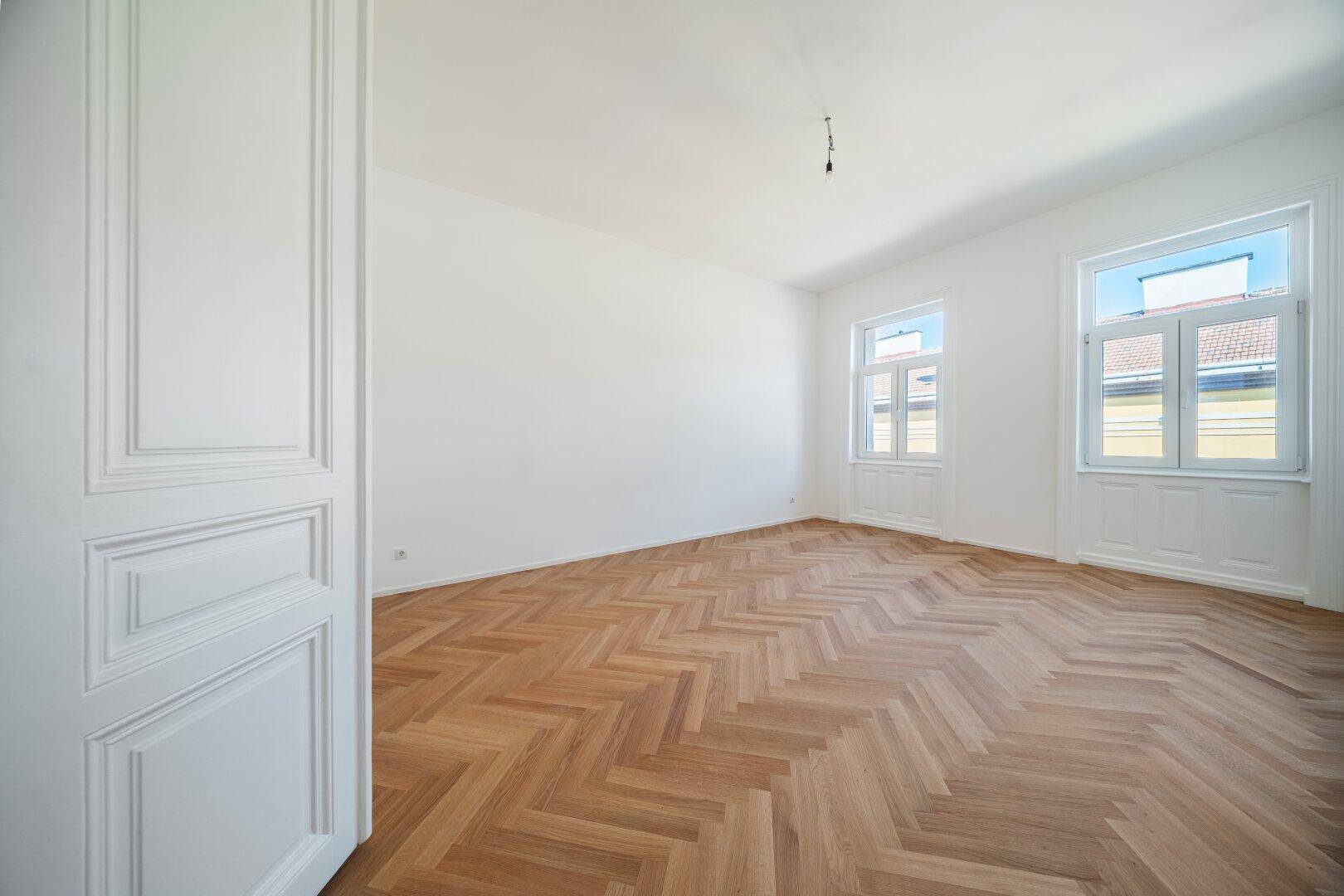 Helles Zimmer mit Fischgrätparkett und zwei Fenstern, klassische Türdetails, frisch gestrichen.