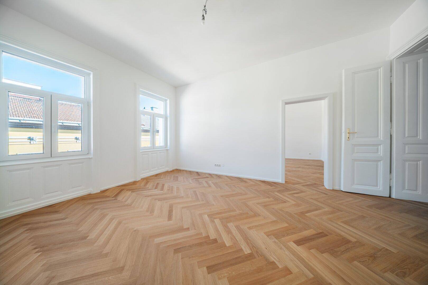 Geräumiges Zimmer mit Fischgrätparkett und mehreren Türen, die zu anderen Bereichen führen.