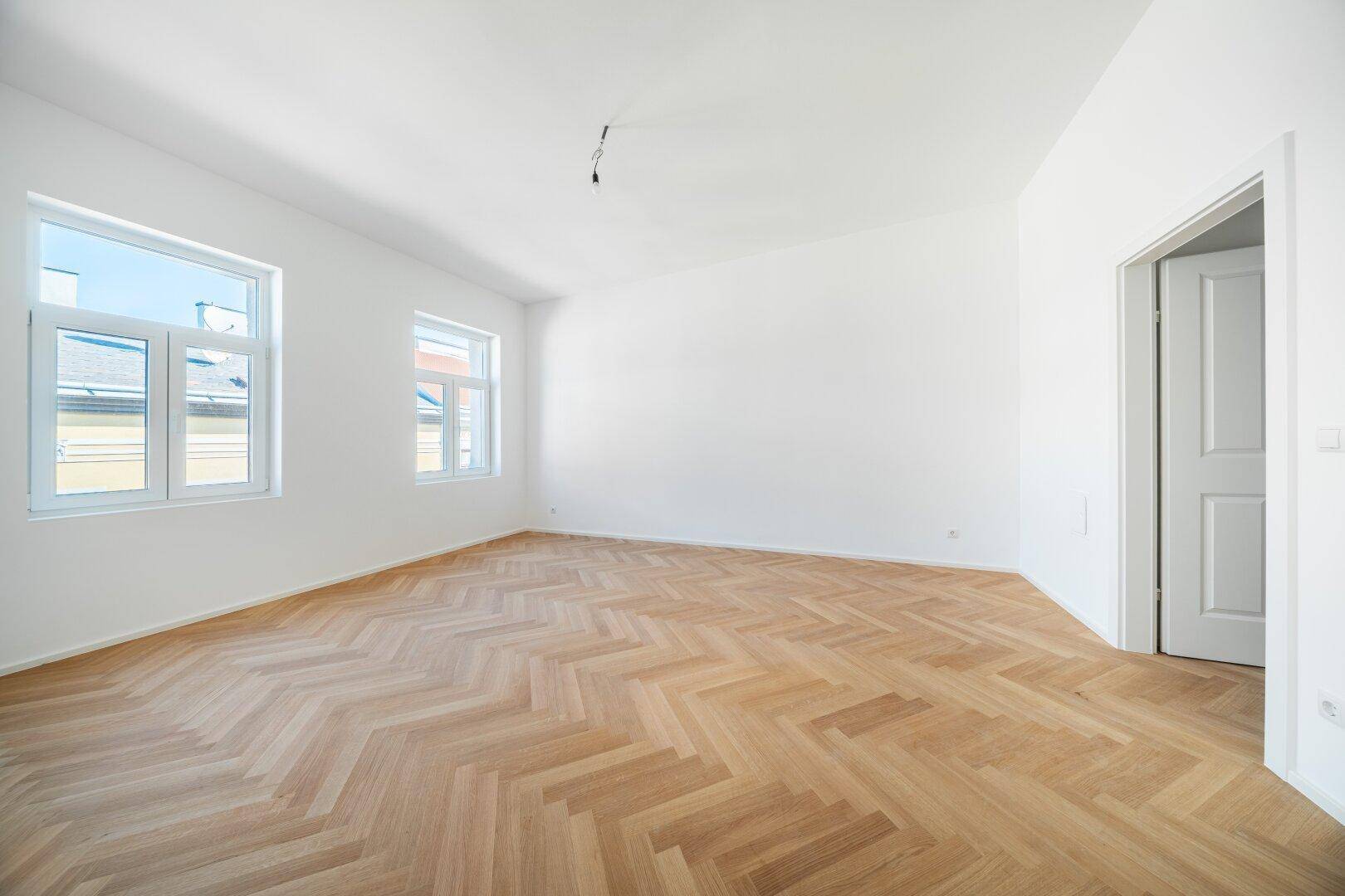 Lichtdurchflutetes Zimmer mit Fischgrätparkett und zwei Fenstern, bereit zum Einrichten.