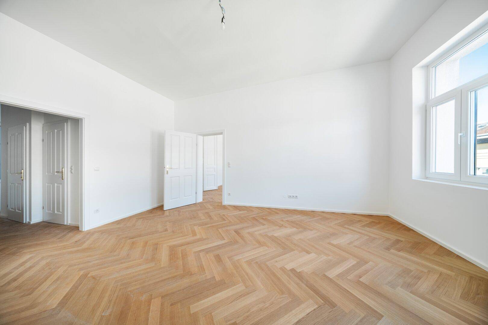 Geräumiges Zimmer mit Fischgrätparkett, einem Fenster und offenen Türen zu angrenzenden Räumen.