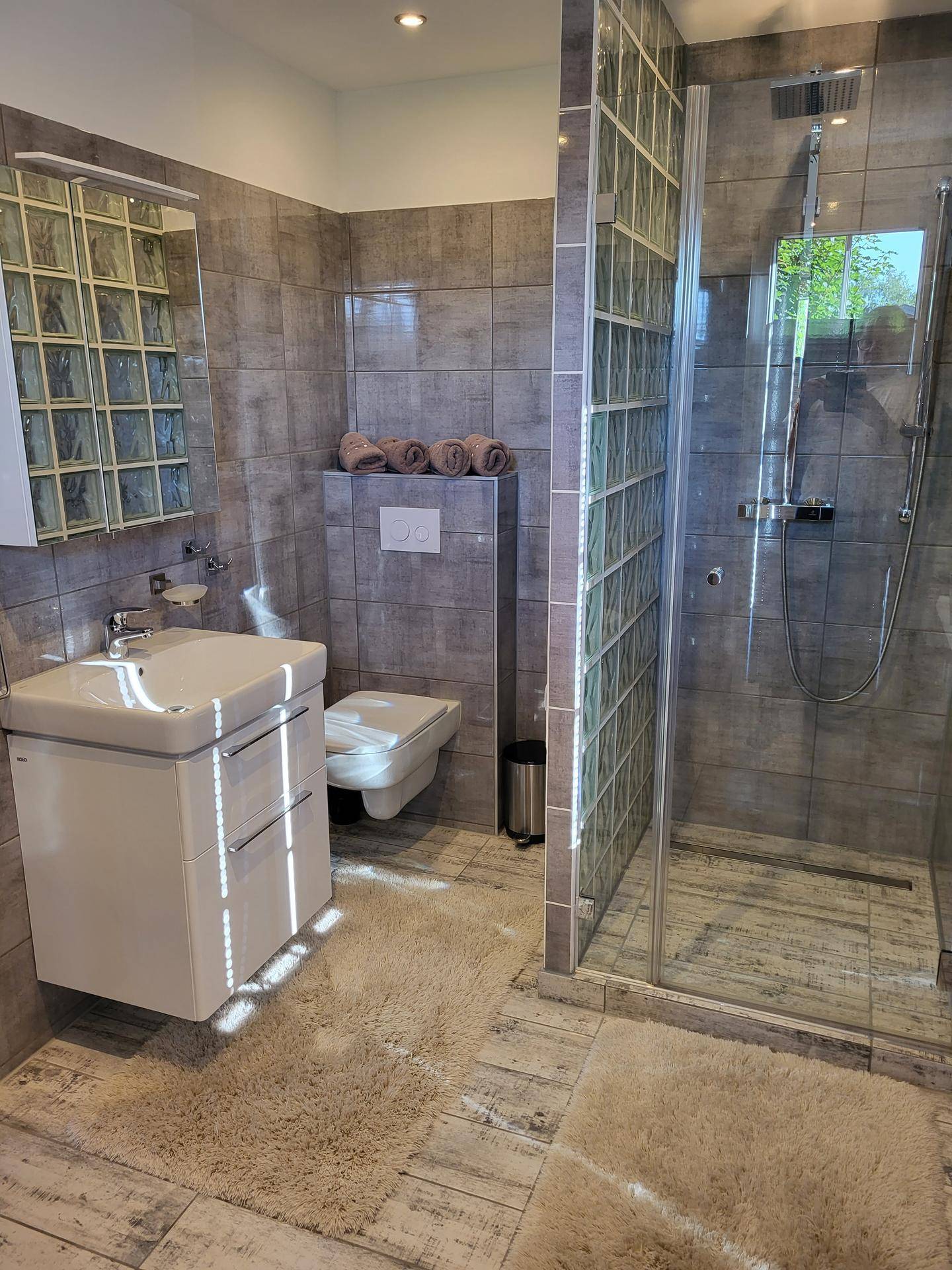 Modernes Badezimmer mit grauen Fliesen, Duschkabine aus Glasbausteinen und Hänge-WC.