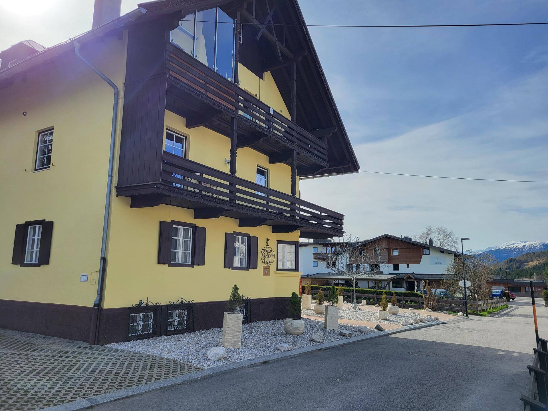 Seitenansicht des gelben Hauses mit Holzbalkonen, großer Glasfront und Bergblick im Hintergrund.