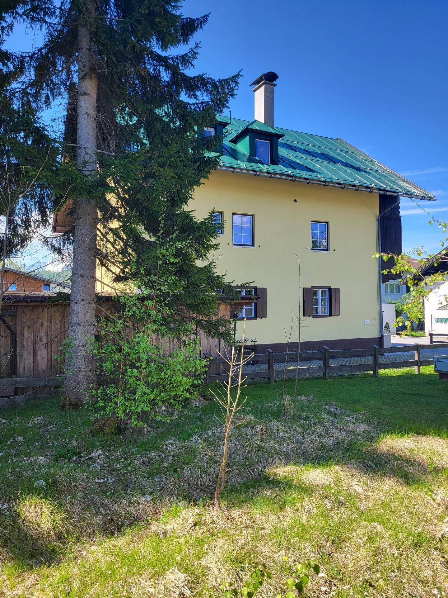Gelbes Haus mit grünem Dach und Schornstein, umgeben von hohen Bäumen und einem gepflegten Rasen.