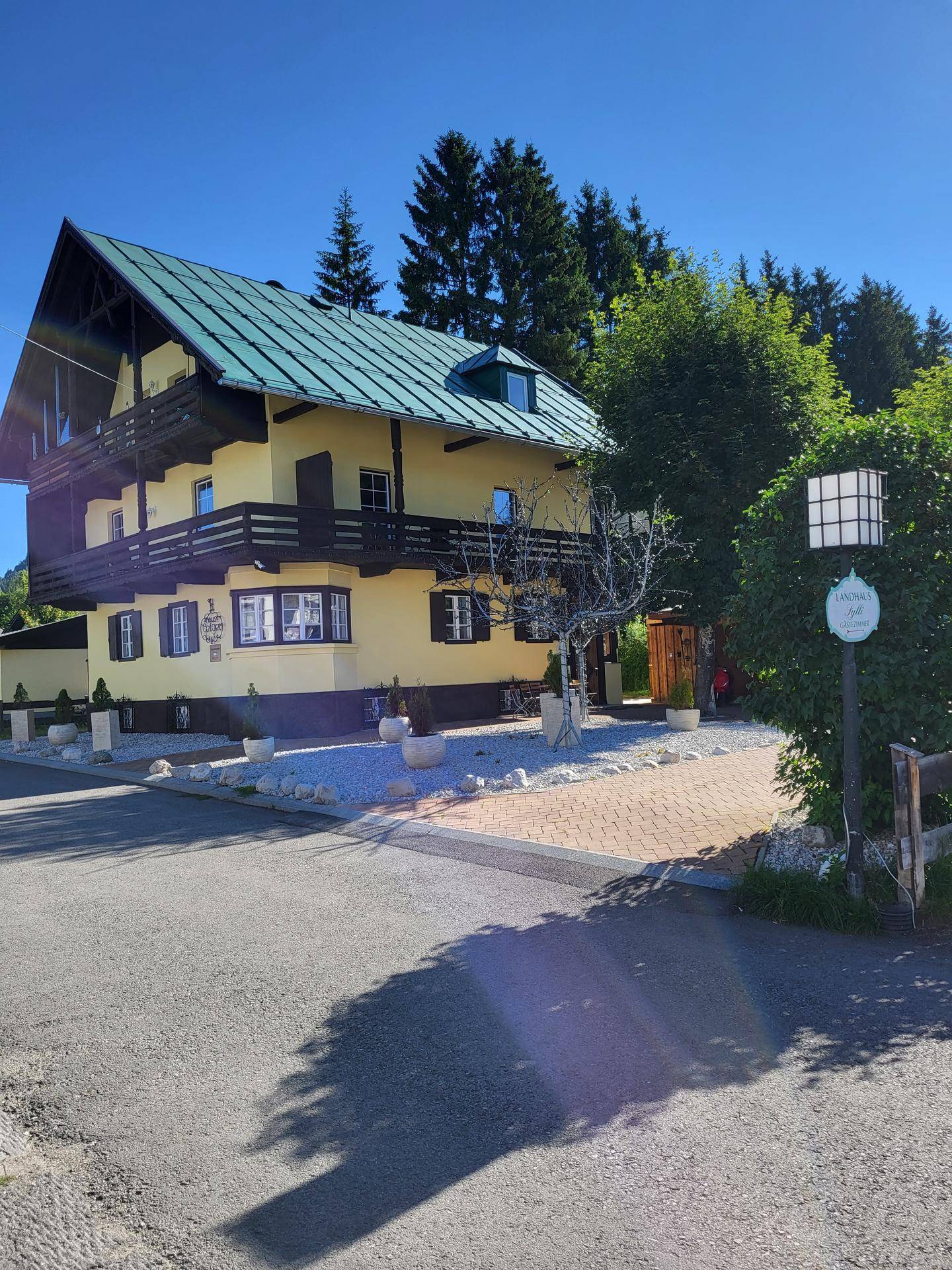 Gelbes Haus mit Holzbalkonen und grünem Dach, umgeben von Bäumen und einer Straße.