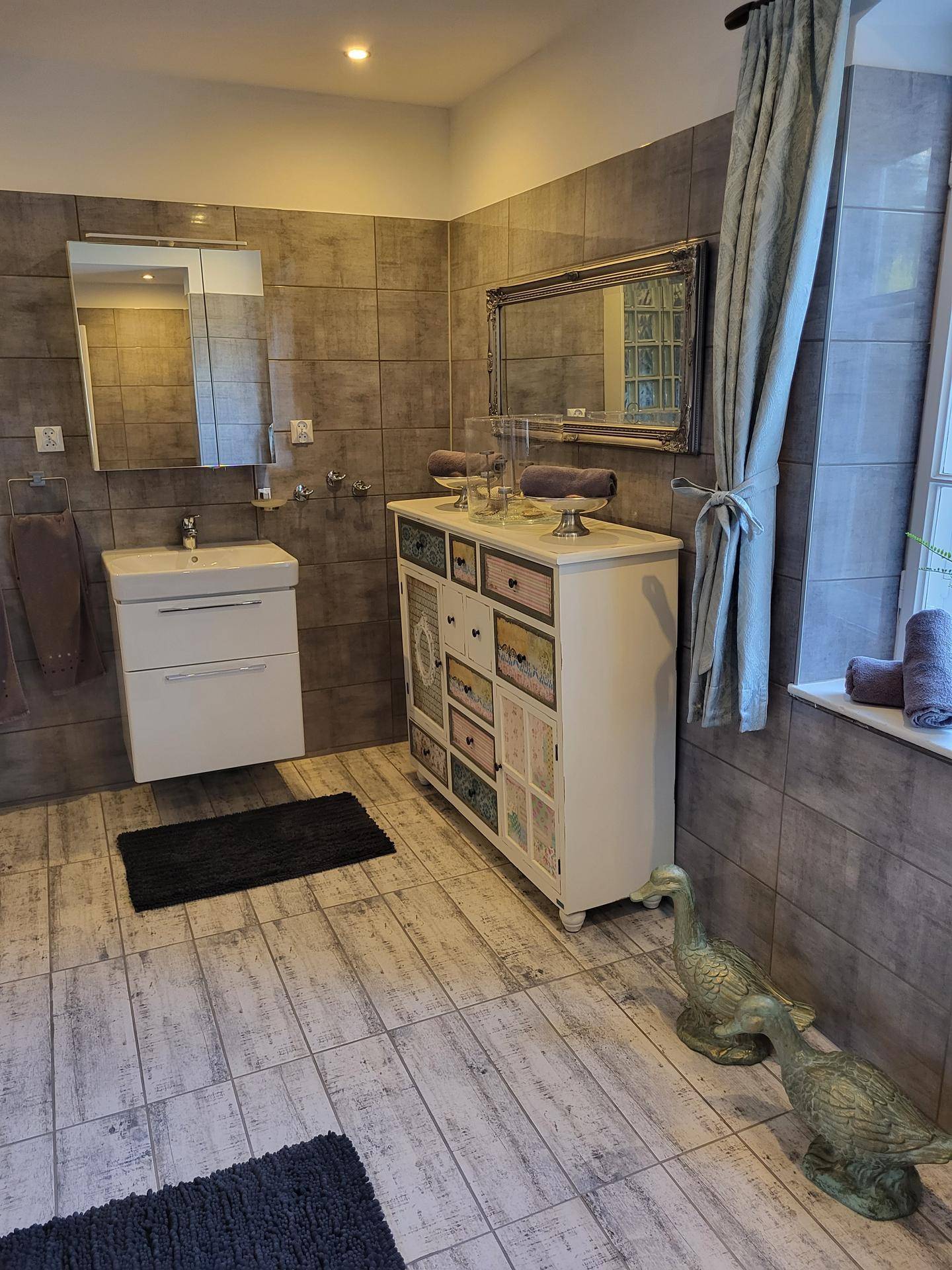 Geräumiges Badezimmer mit grauen Fliesen, Waschtisch und einem dekorativen Spiegelschrank.