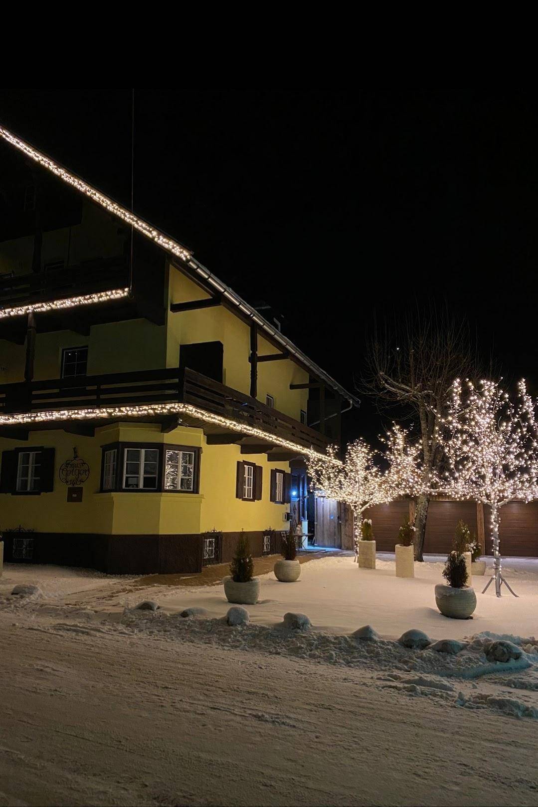 Gelbes Haus bei Nacht, festlich beleuchtet mit Lichterketten und Schnee im Vorgarten.