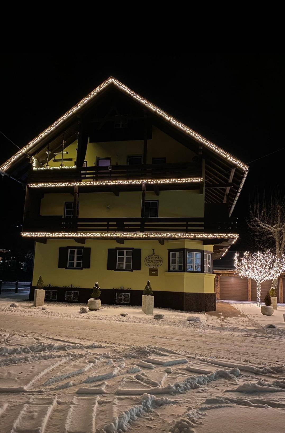 Winterliche Nachtansicht des Hauses mit festlicher Beleuchtung an Balkonen und Dach.