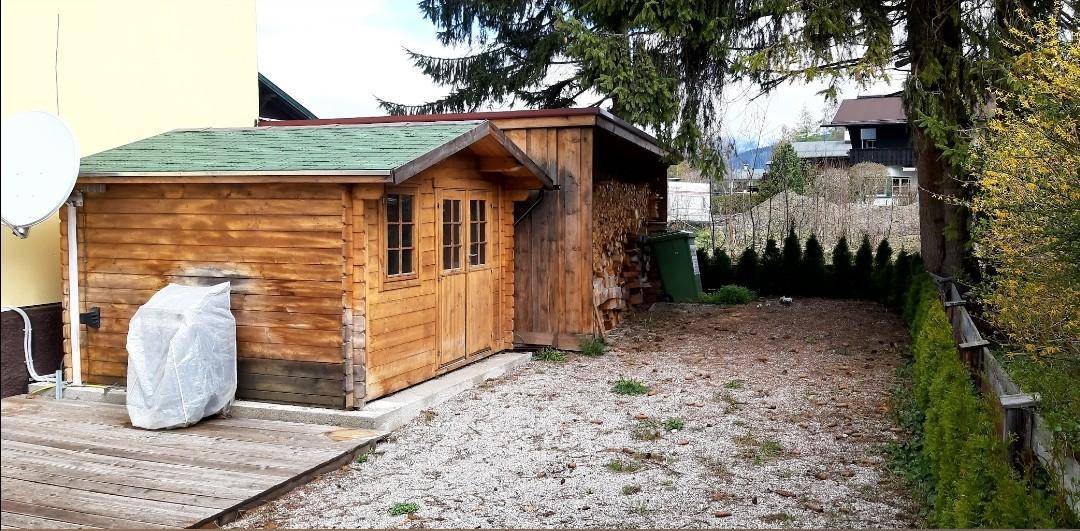 Hinterhofbereich mit Holzschuppen, Holzstapel und einer Hecke, umgeben von Bäumen.