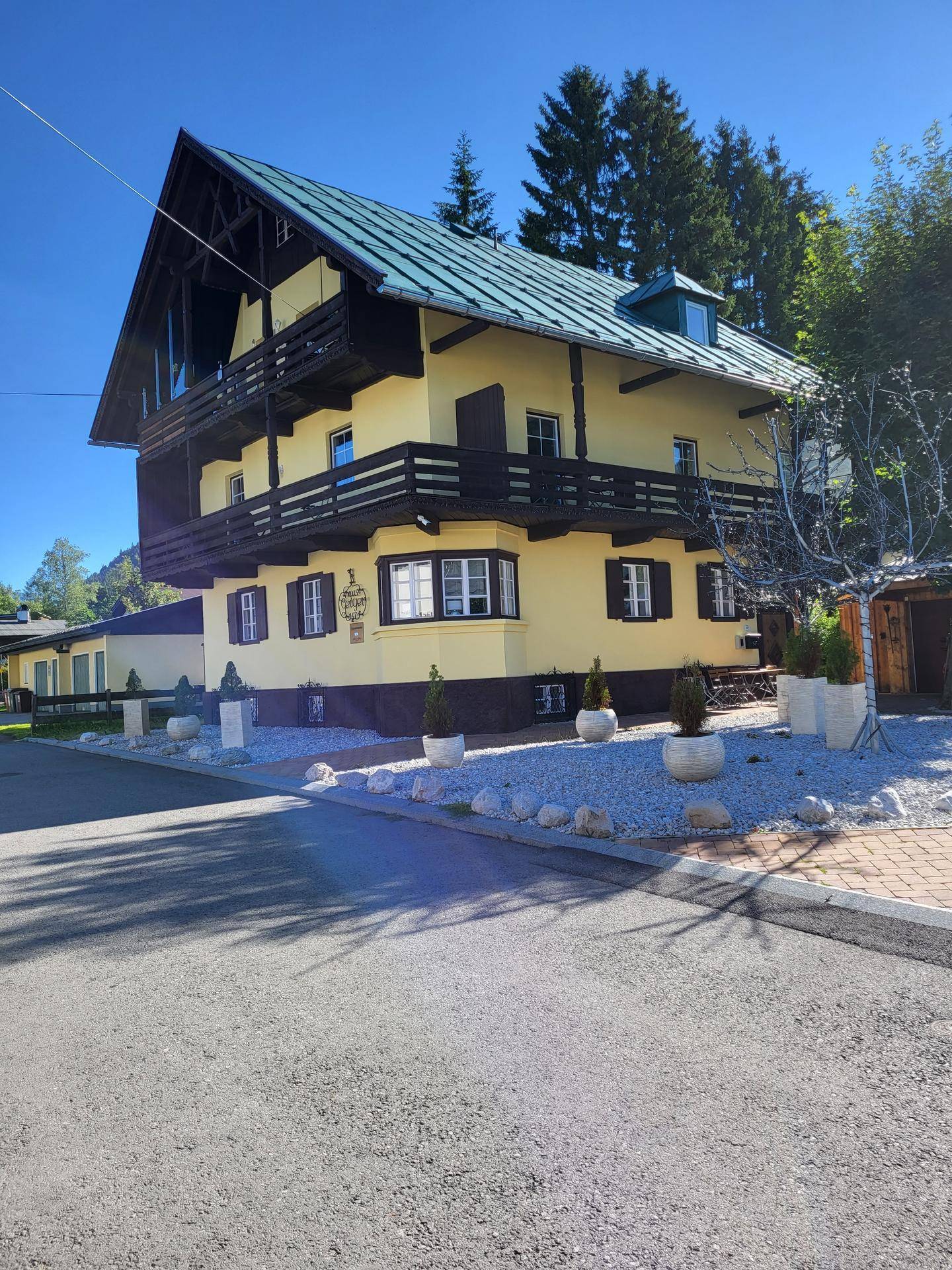 Sonnenbeschienenes gelbes Haus mit Holzbalkonen und grünem Dach in alpiner Umgebung.