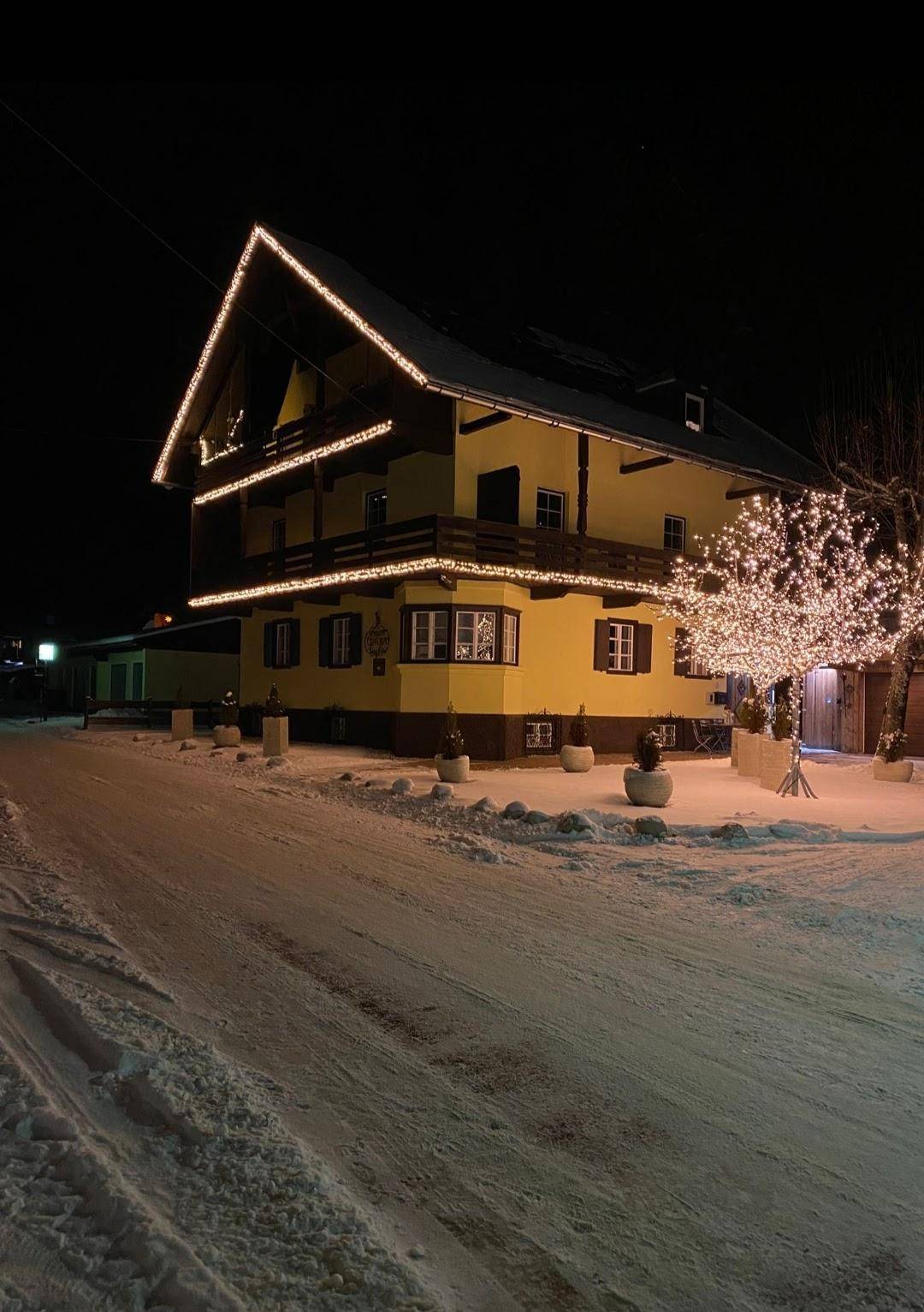 Traditionelles gelbes Haus im Winter, festlich beleuchtet mit Lichterketten und verschneiter Umgebung.