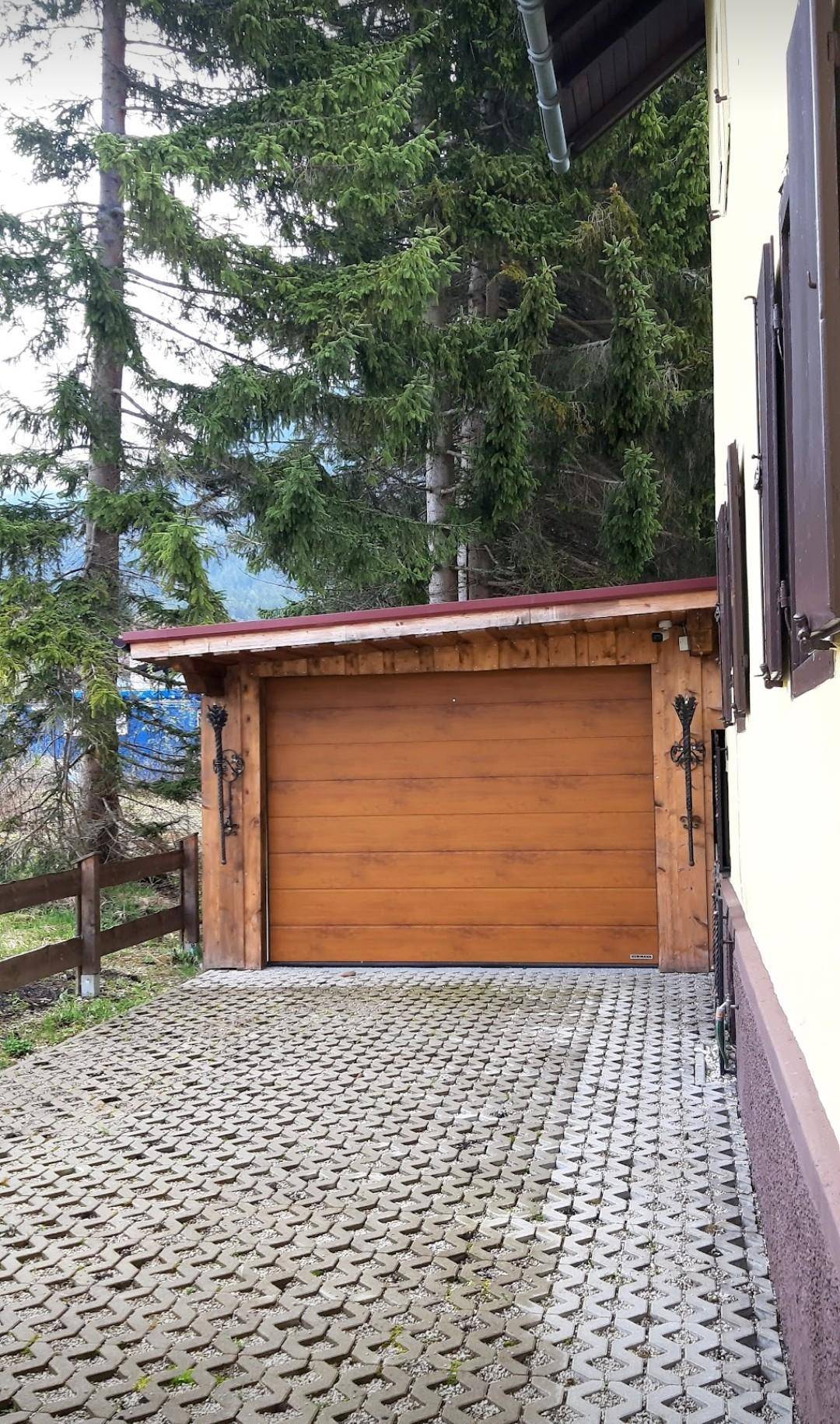 Einzelne Garage mit Holztor und gepflasterter Zufahrt, umgeben von hohen Nadelbäumen.