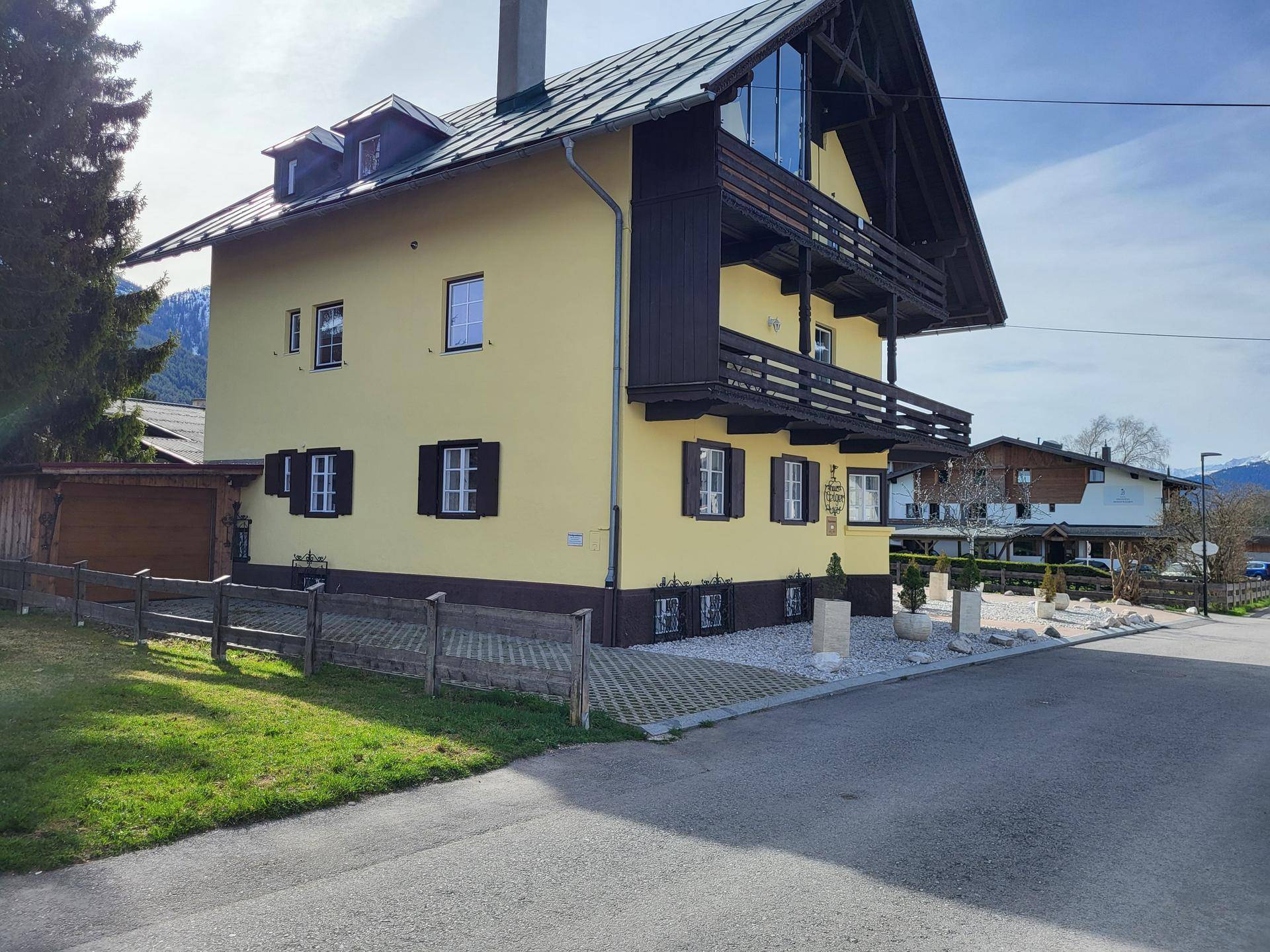 Seitenansicht des gelben Hauses mit Holzbalkonen, großer Glasfront und Bergblick im Hintergrund.