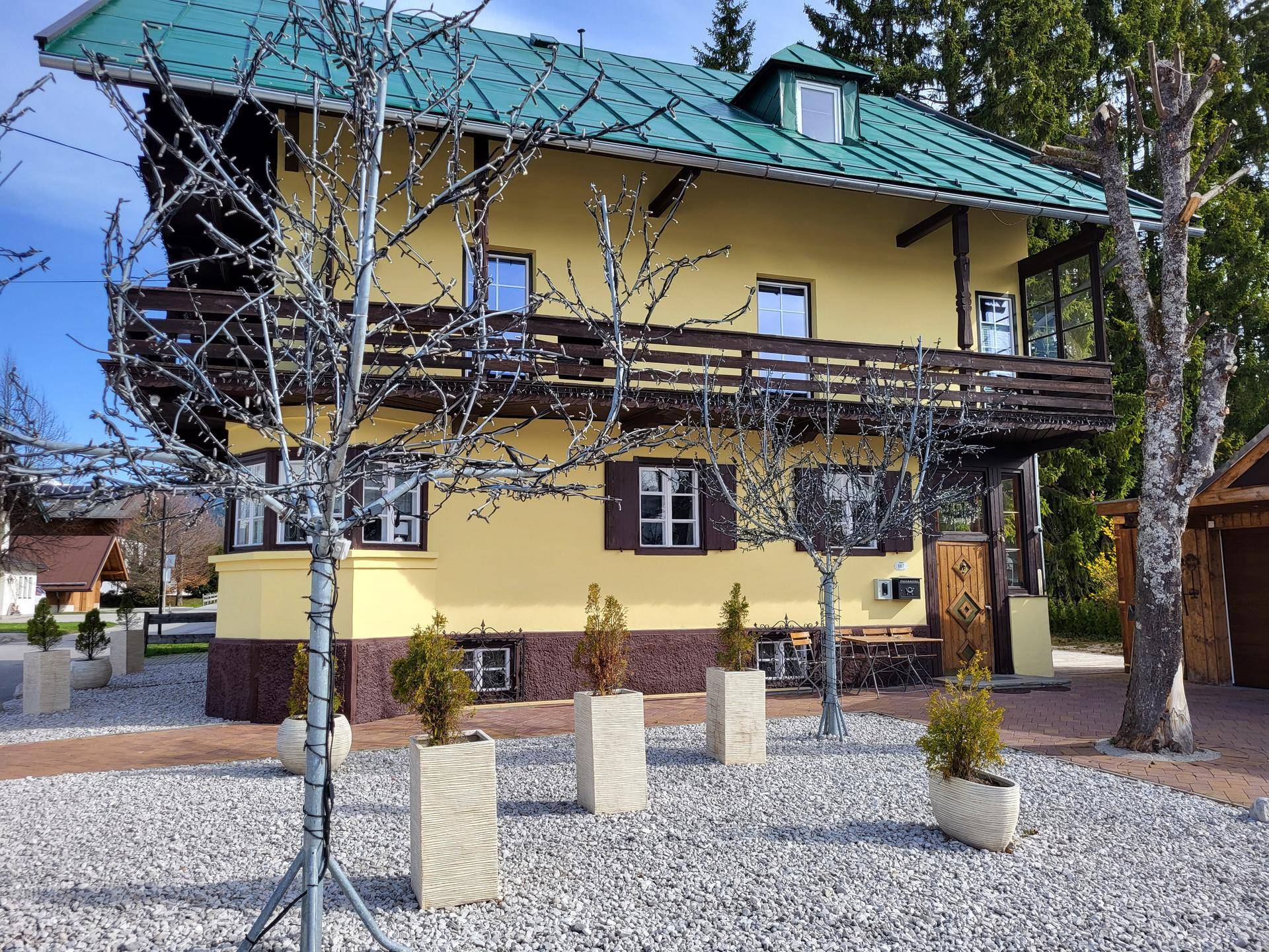 Gelbes Haus mit grünem Dach, Holzbalkonen und einem Vorgarten mit Zierbäumen.