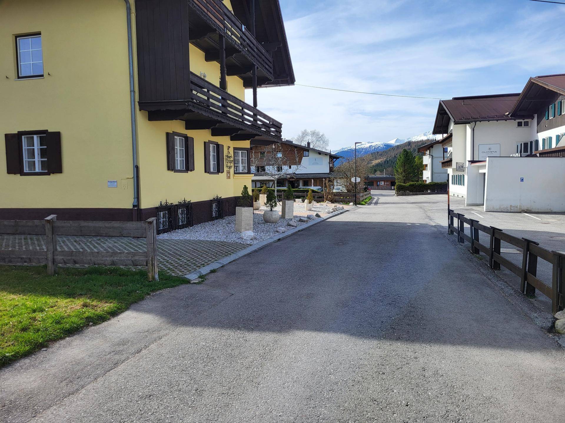 Gelbes Haus mit Holzbalkonen an einer Straße, umgeben von weiteren Gebäuden und Bergen im Hintergrund.
