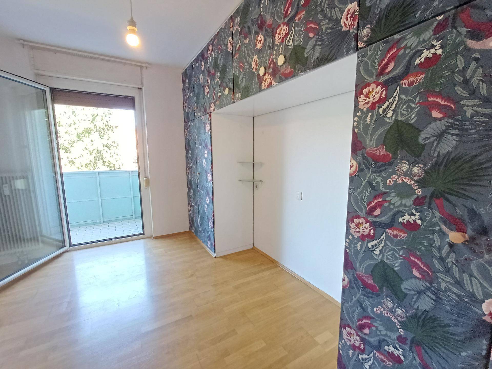 Geräumiges Zimmer mit Parkettboden und auffälligen floralen Einbauschränken sowie Balkonzugang.