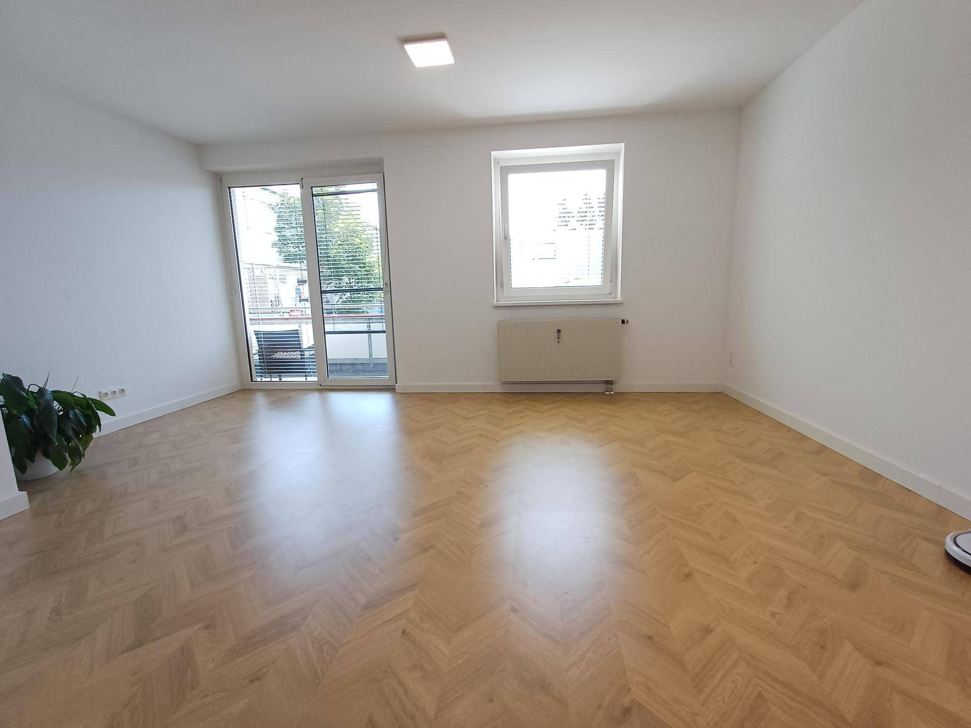 Geräumiges, leeres Wohnzimmer mit hellem Fischgrätparkett und großem Fenster, bietet viel Tageslicht.