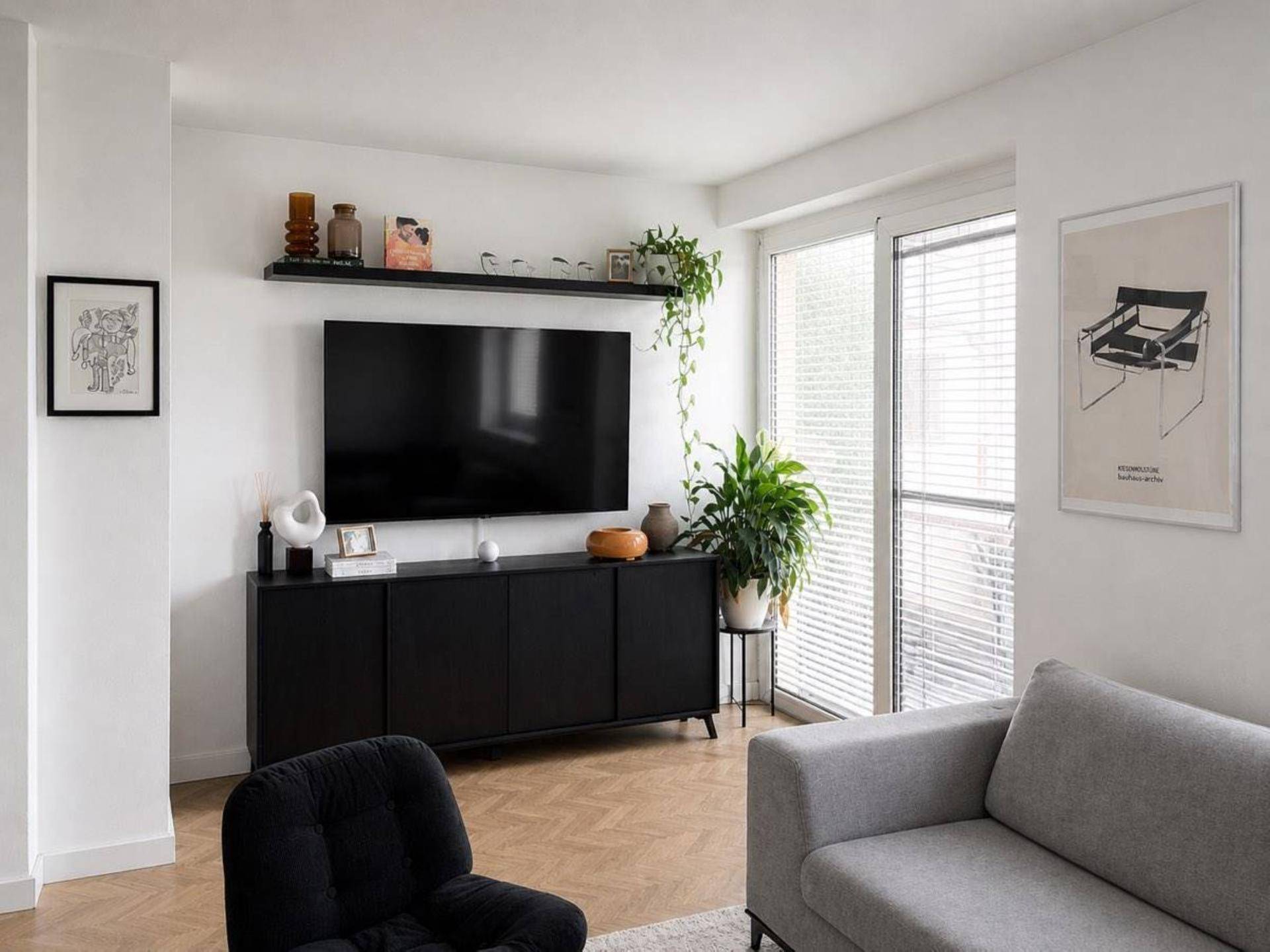 Modern eingerichtetes Wohnzimmer mit Fernseher, Sideboard und Pflanzen, hell durch große Fenster.