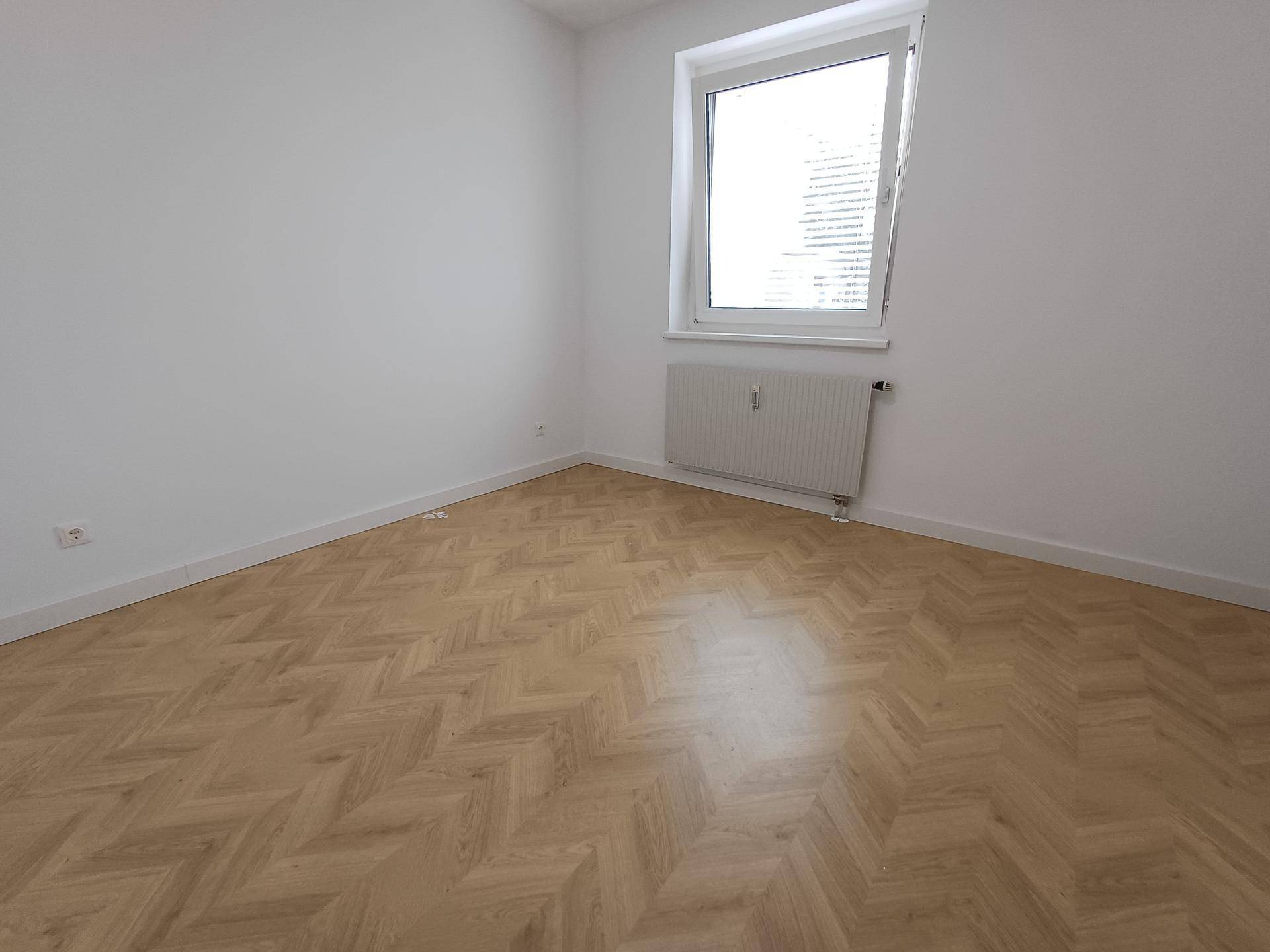 Leeres Schlafzimmer mit hellem Fischgrätparkett und großem Fenster, bietet viel Tageslicht.