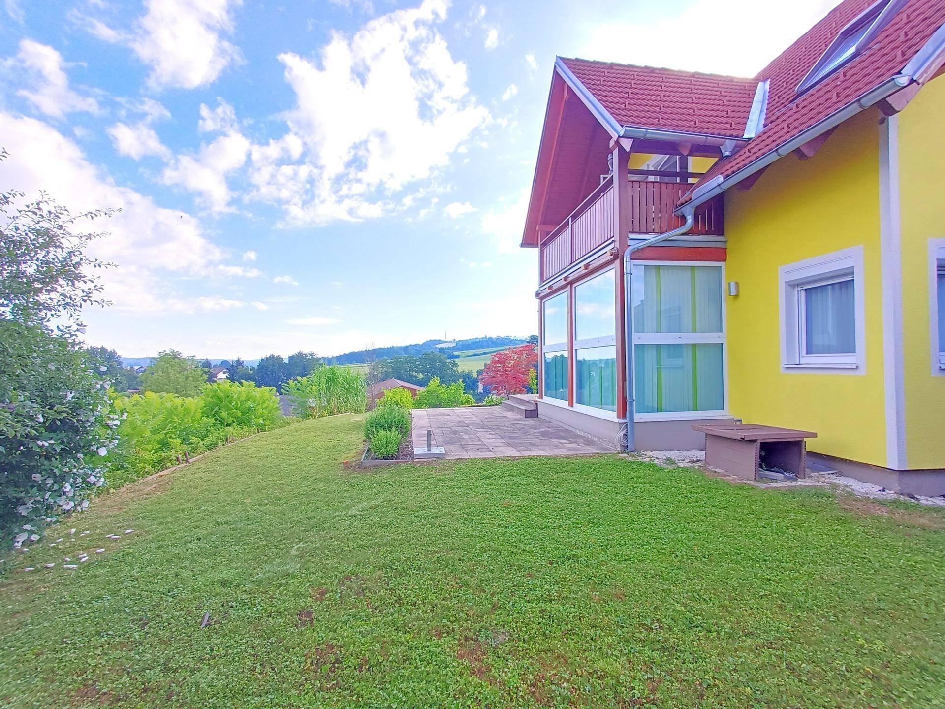 Gelbes Einfamilienhaus mit rotem Dach, Terrasse und Balkon, umgeben von grüner Natur.