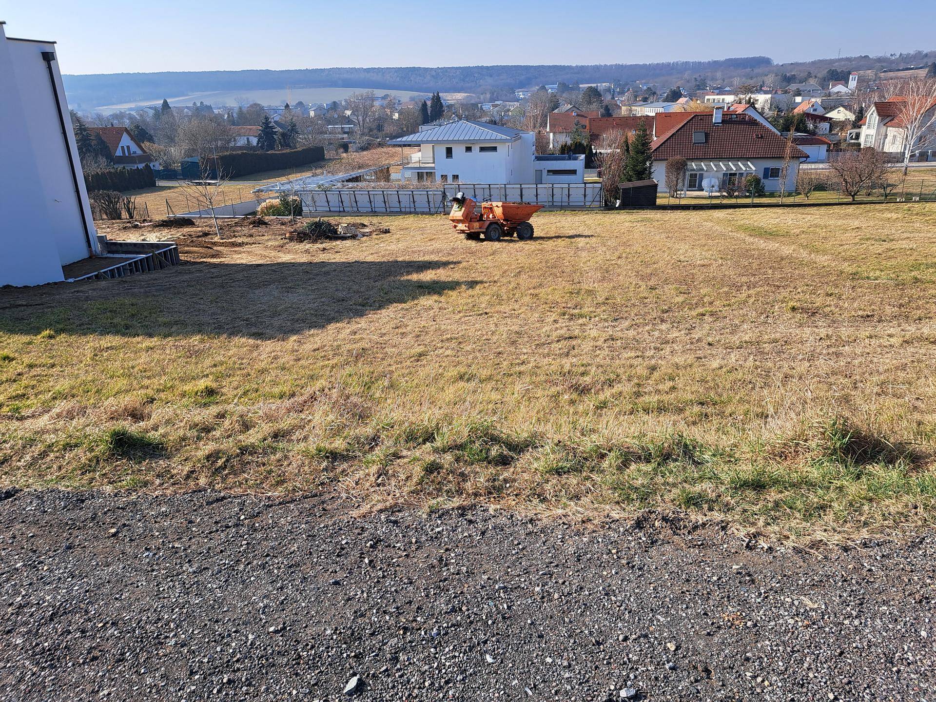 Grundstück mit trockenem Gras, einem kleinen Baufahrzeug und Häusern in der Ferne, mit Schotter im Vordergrund.